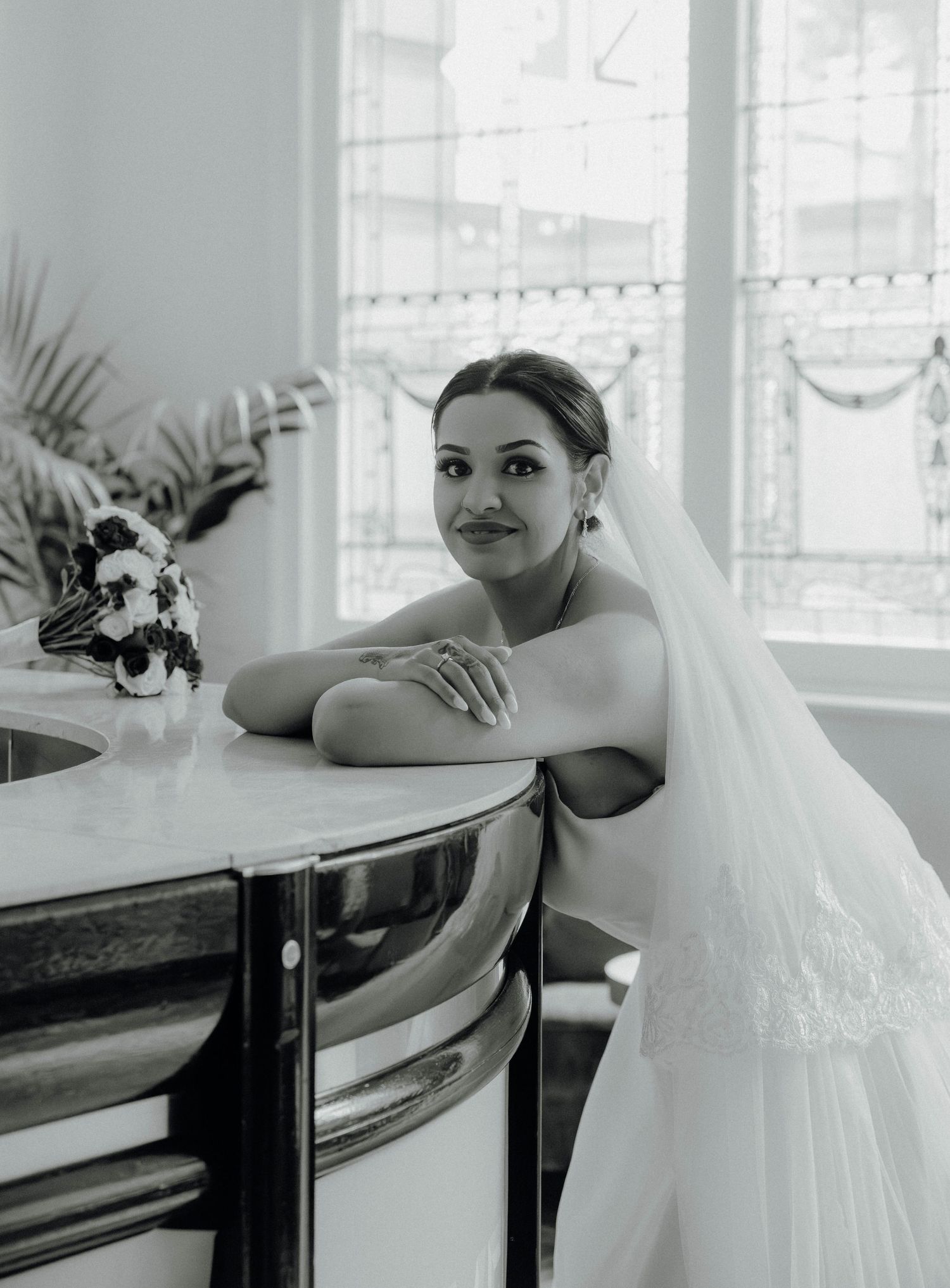 Black and white photo of a smiling bride in a white gown and veil seated at a table with a bouquet nearby.