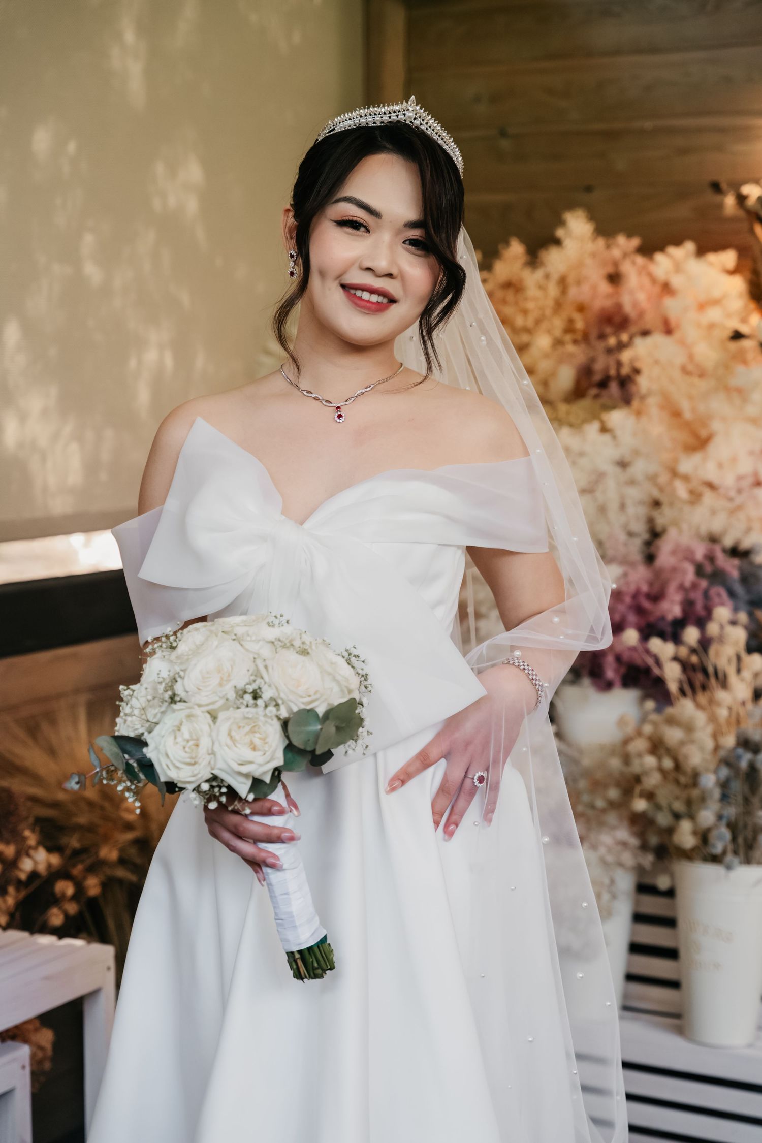 Bride in white off-shoulder wedding dress holding white rose bouquet with floral decor and dreamy backdrop.