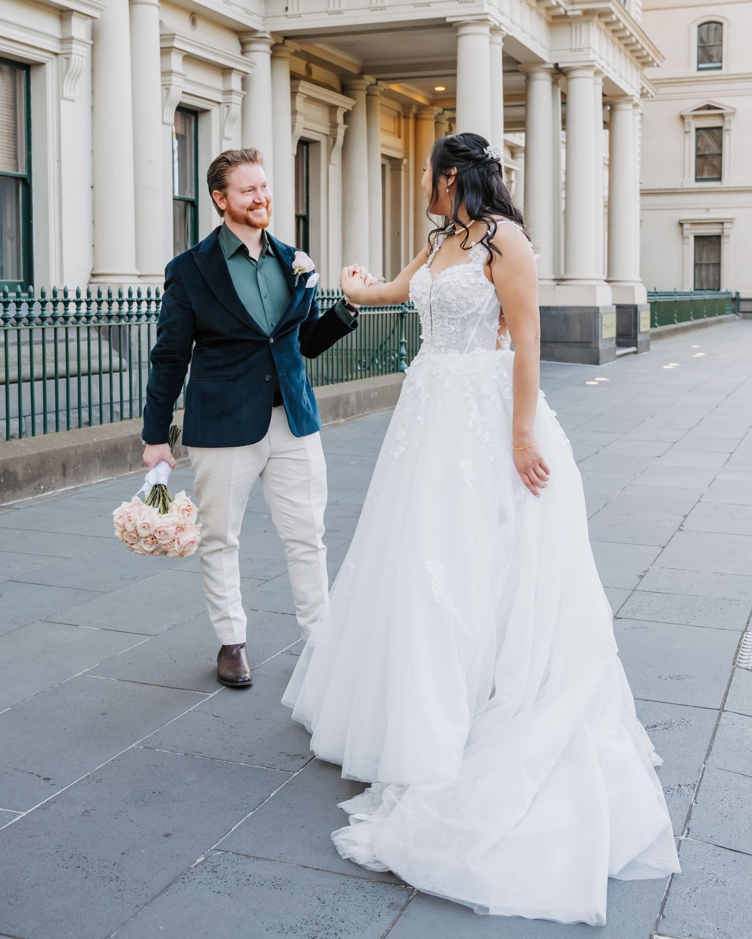 A lively wedding couple shares a joyful moment while walking together outside a grand historic building with white columns.