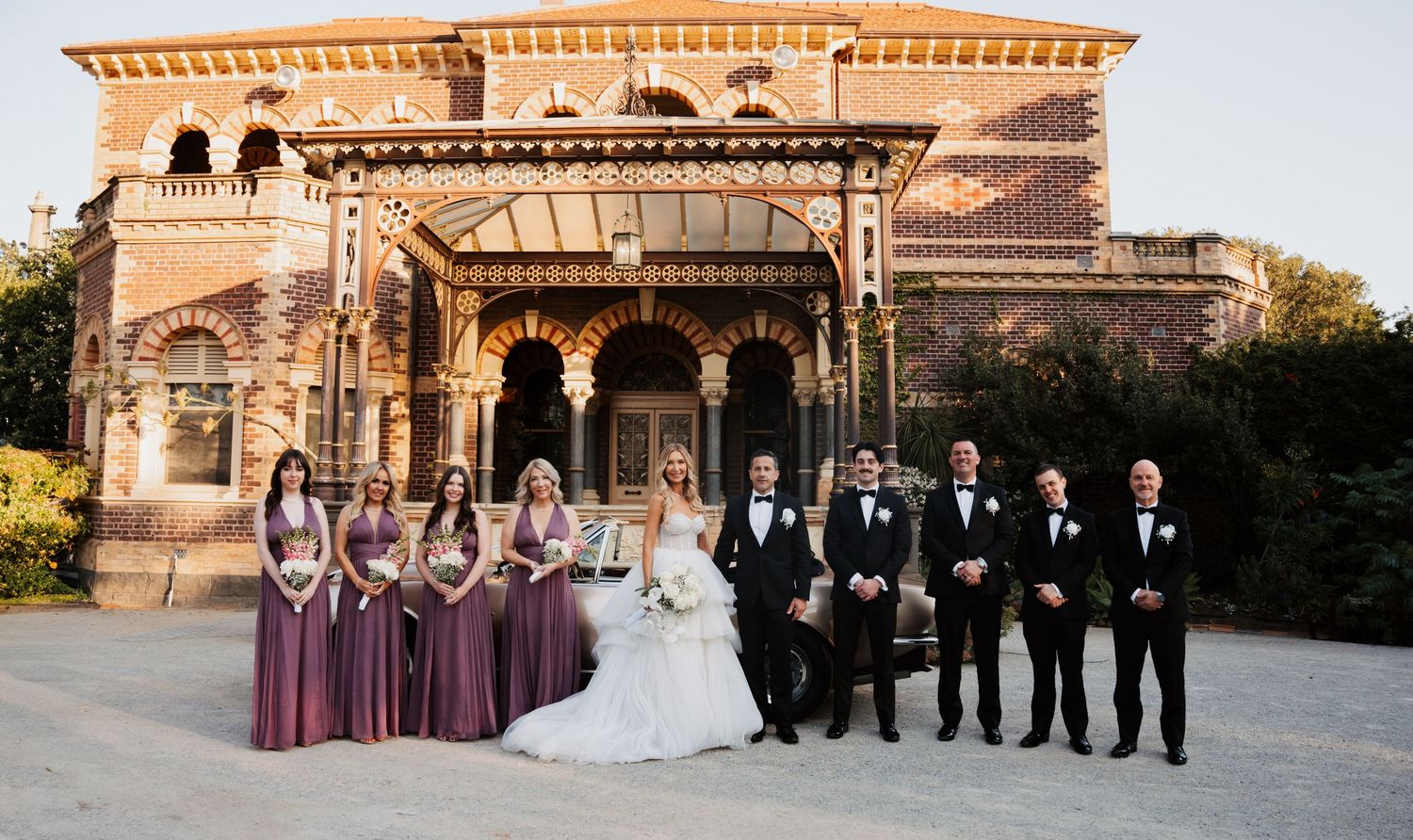 Wedding party poses in front of ornate Victorian mansion with elegant architectural details at sunset.