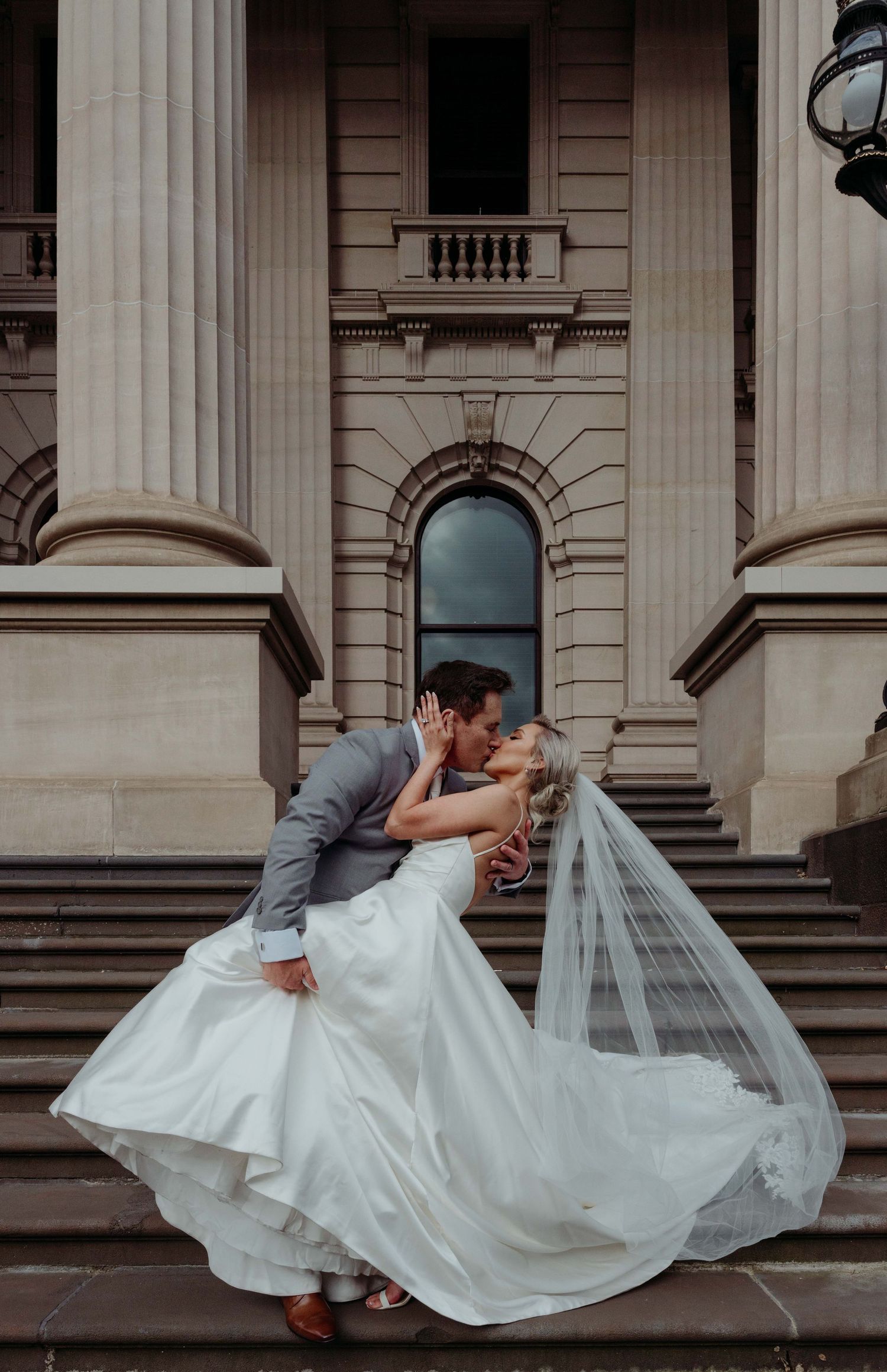 A romantic wedding couple embraces on grand stone steps of a historic building with flowing white veil and classic architecture.