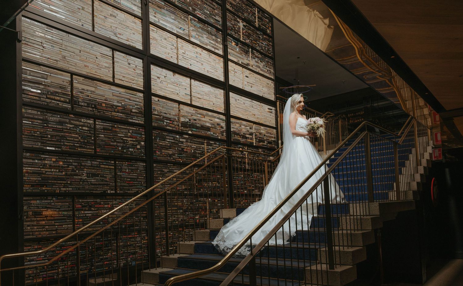 A bride in a flowing white gown stands elegantly on a rustic wooden staircase with exposed brick walls.