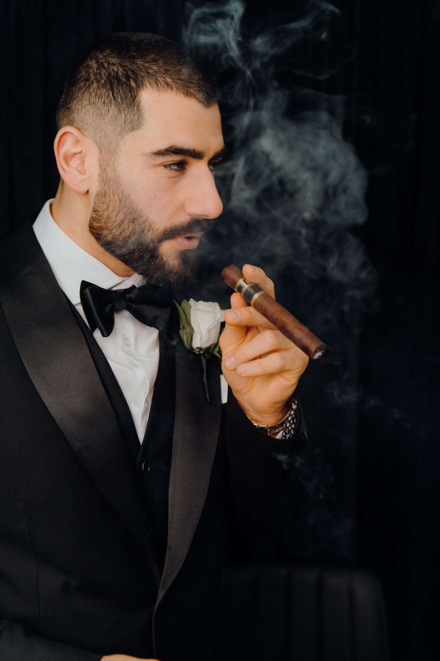 A person in a black tuxedo smoking a cigar against a dark background with dramatic lighting and smoke effects.