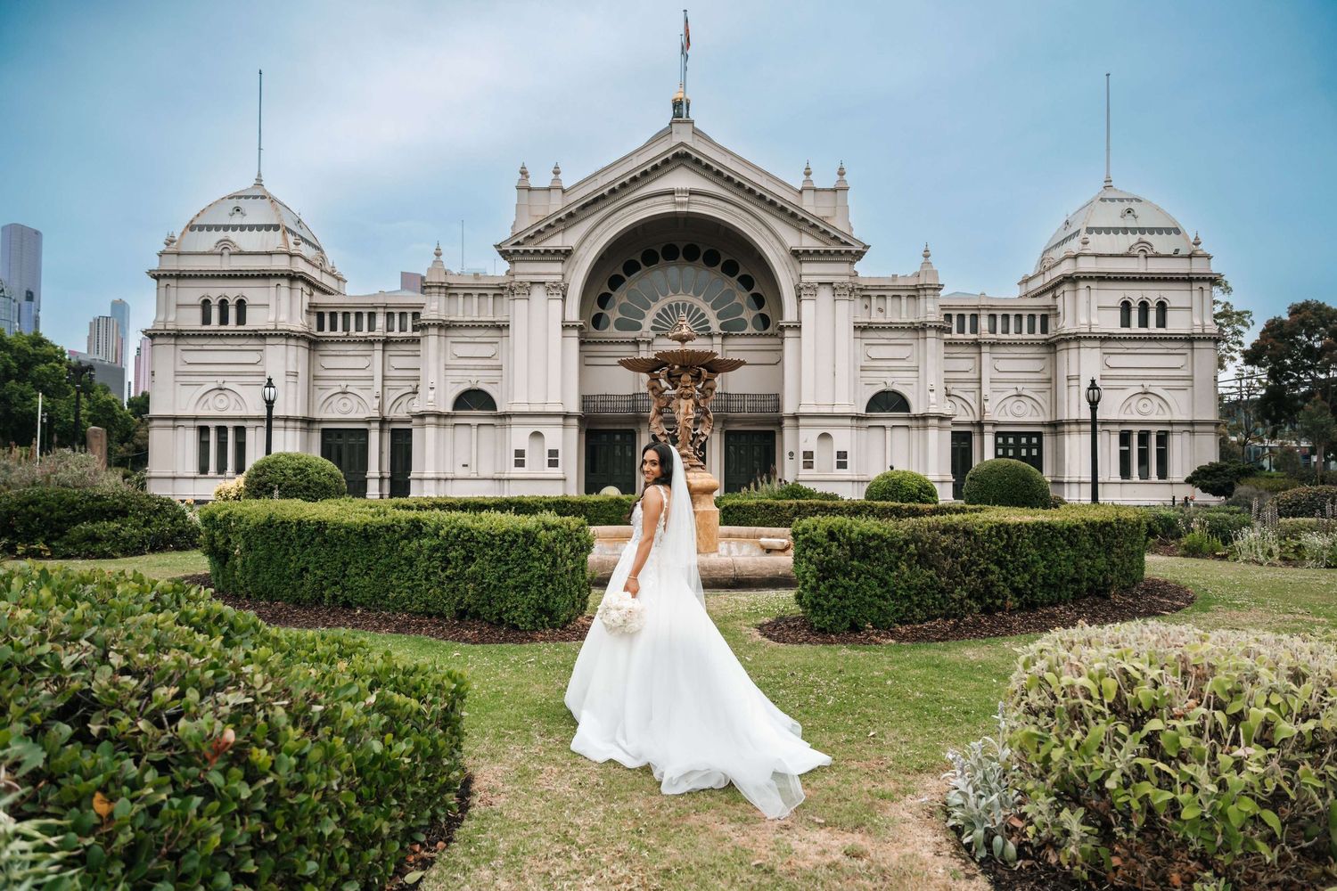 White wedding dress twirls in front of the ornate Royal Exhibition Building in Melbourne with manicured gardens.