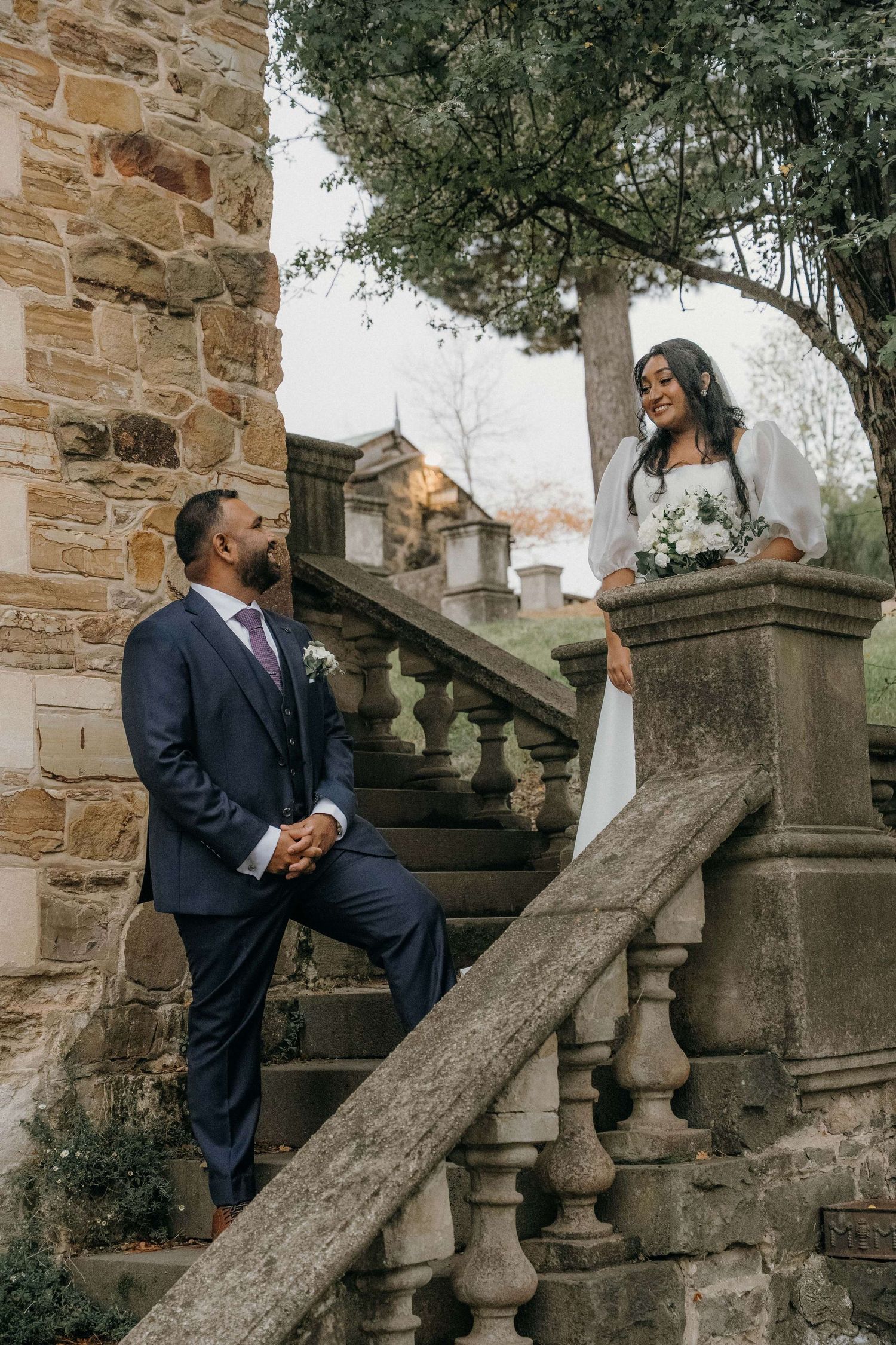 A romantic couple embraces on rustic stone stairs surrounded by lush greenery in an elegant outdoor setting.