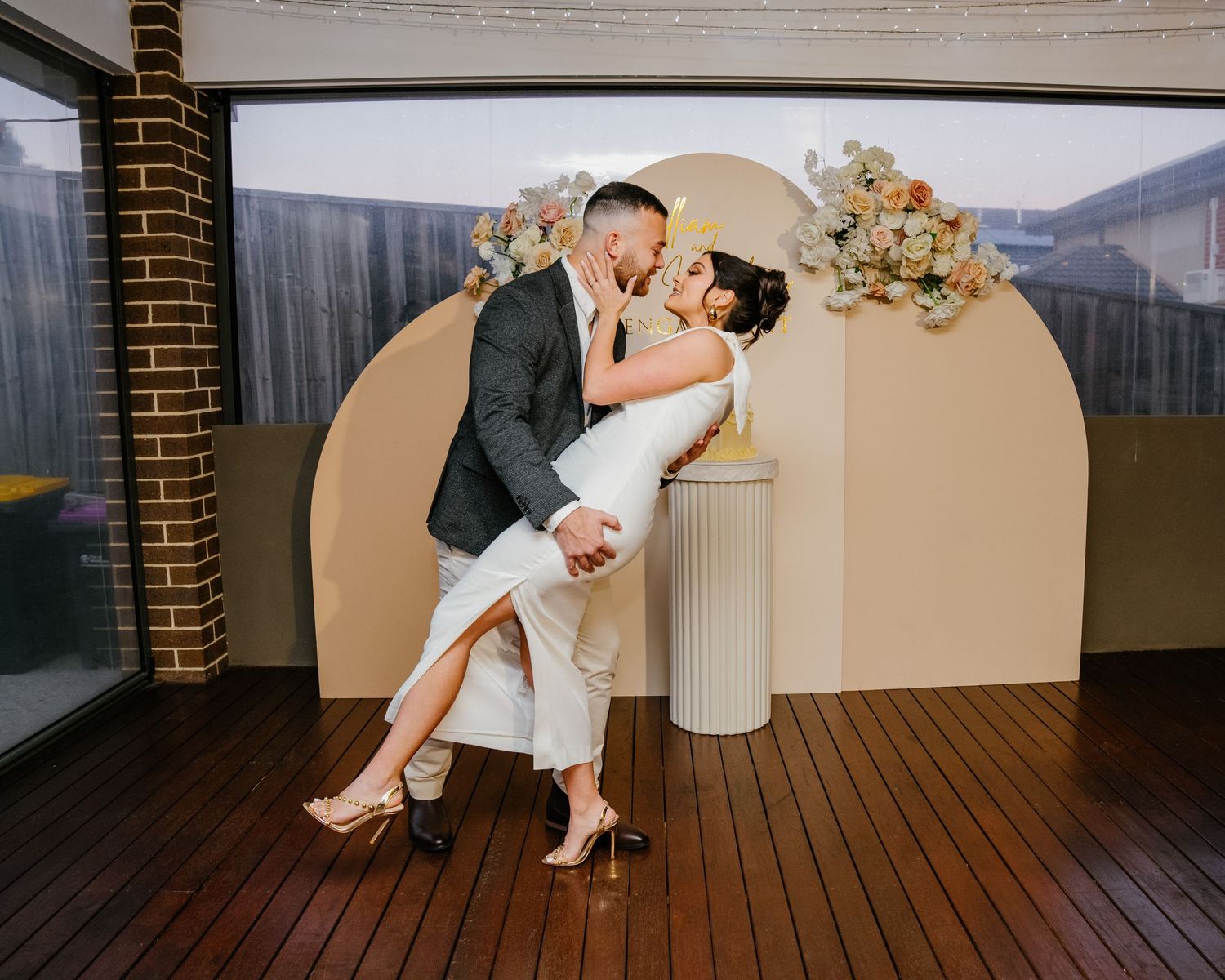 A couple shares a romantic dip pose during their wedding ceremony in front of a modern beige arch and floral decor.