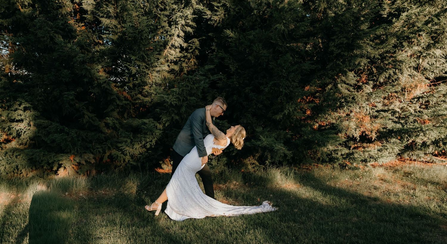 A romantic dip pose between a couple in formal attire against a backdrop of lush green trees and sunlit forest.