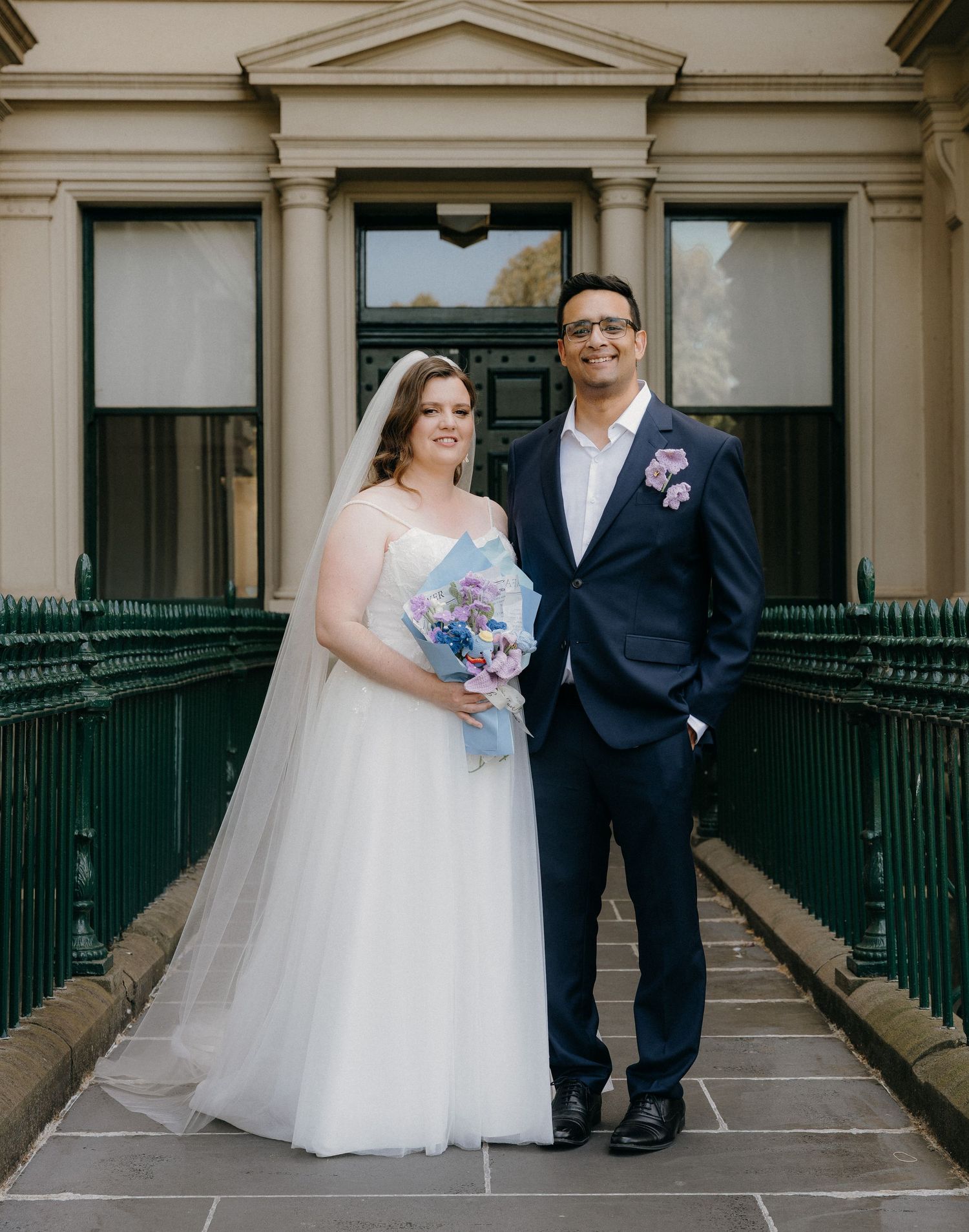 Wedding couple stands on ornate stone steps with white railings in front of a historic building entrance.