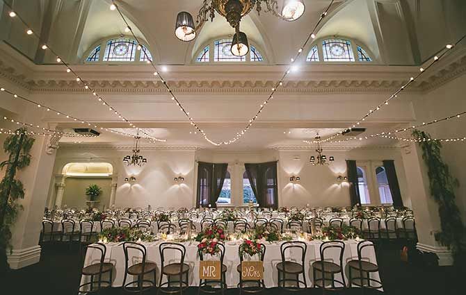 Elegant wedding reception hall with vaulted ceilings, crystal chandeliers and string lights illuminating long dining tables.