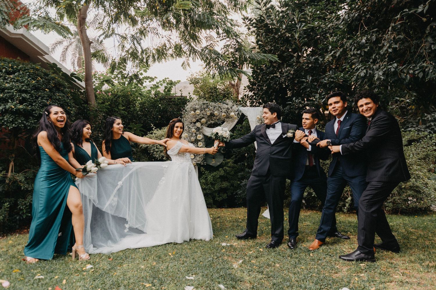 Wedding party poses playfully on garden lawn during outdoor celebration.