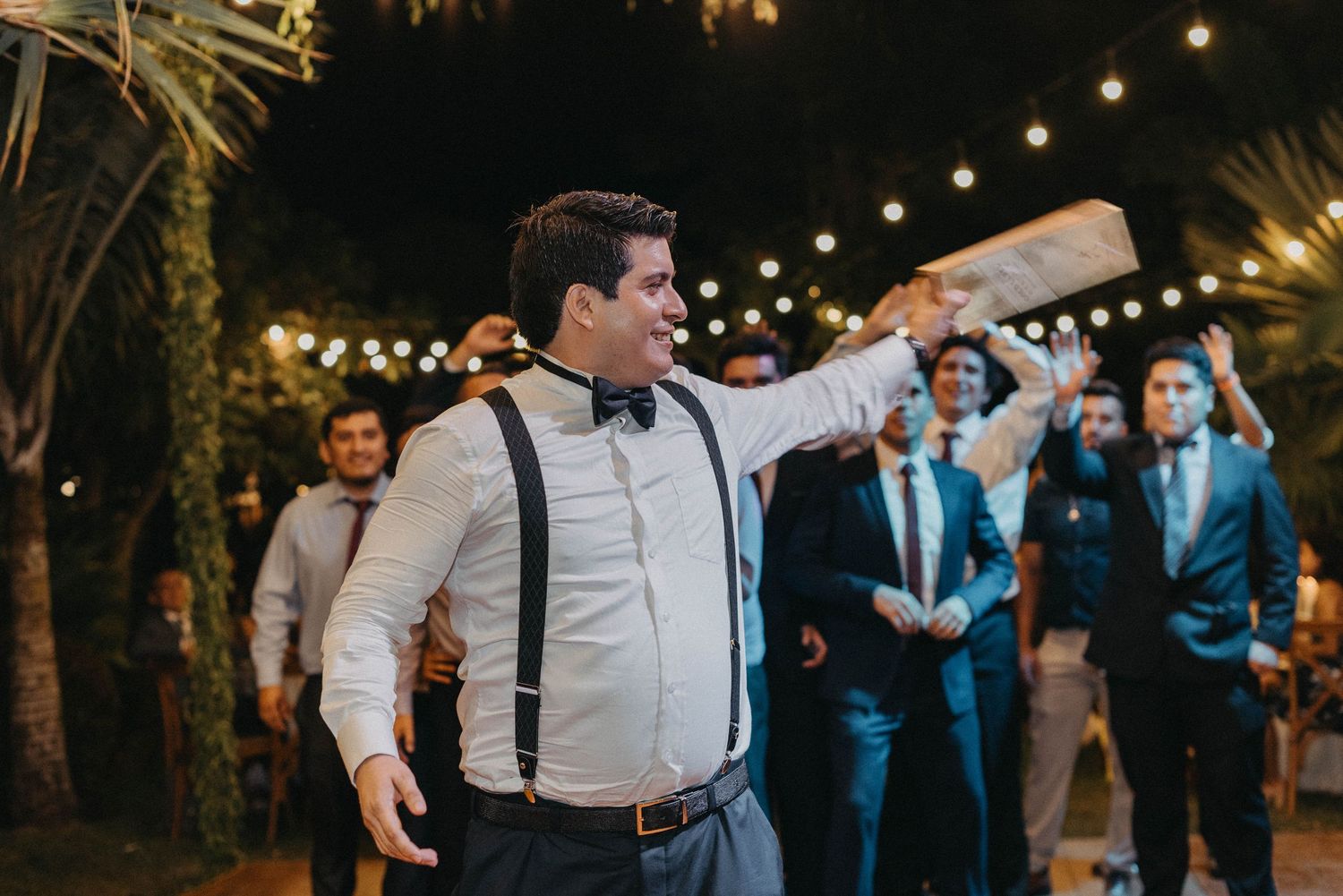Wedding party dances outdoors under string lights at an evening reception celebration.