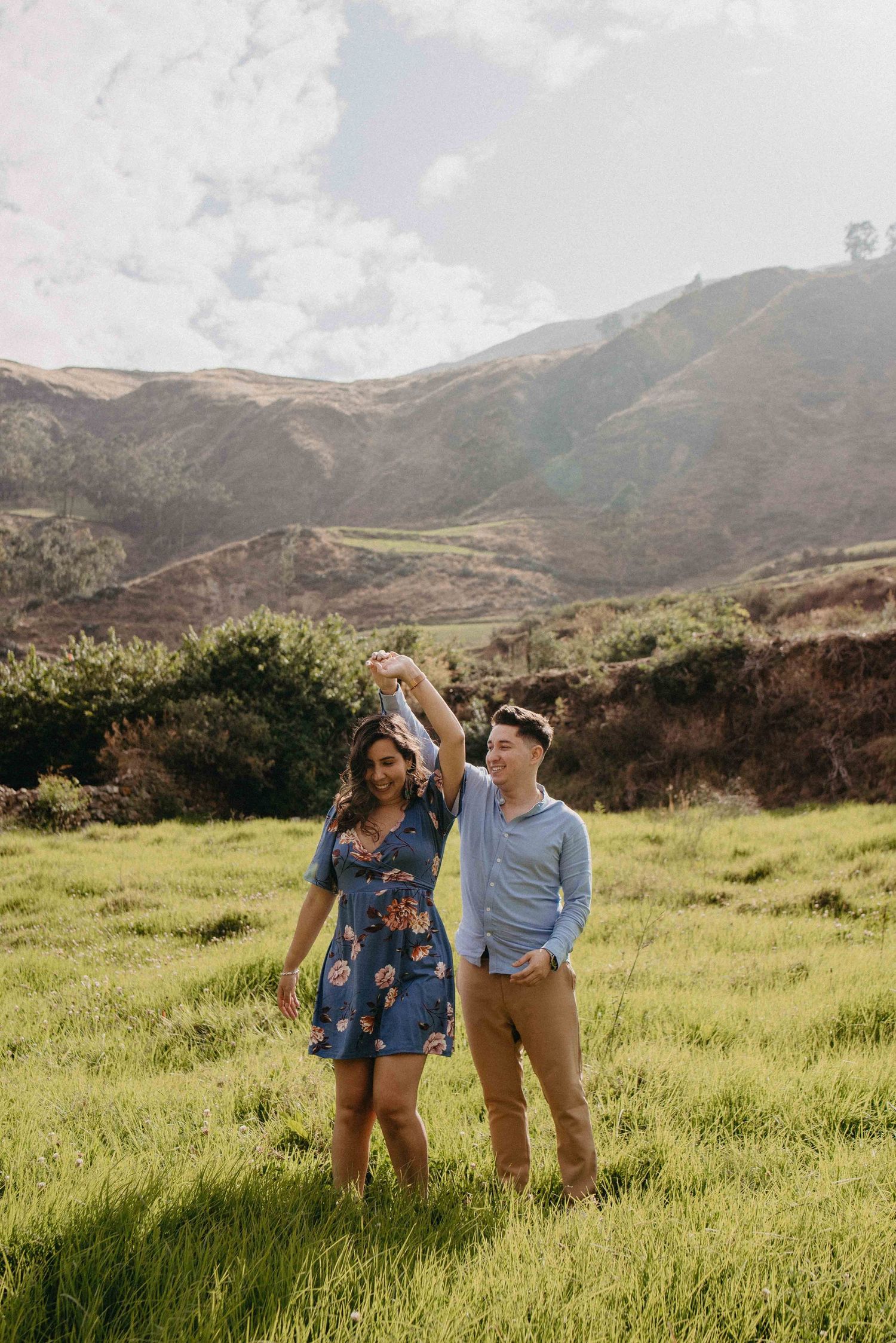 A couple sharing a playful moment in a sunlit meadow with rolling hills behind them.