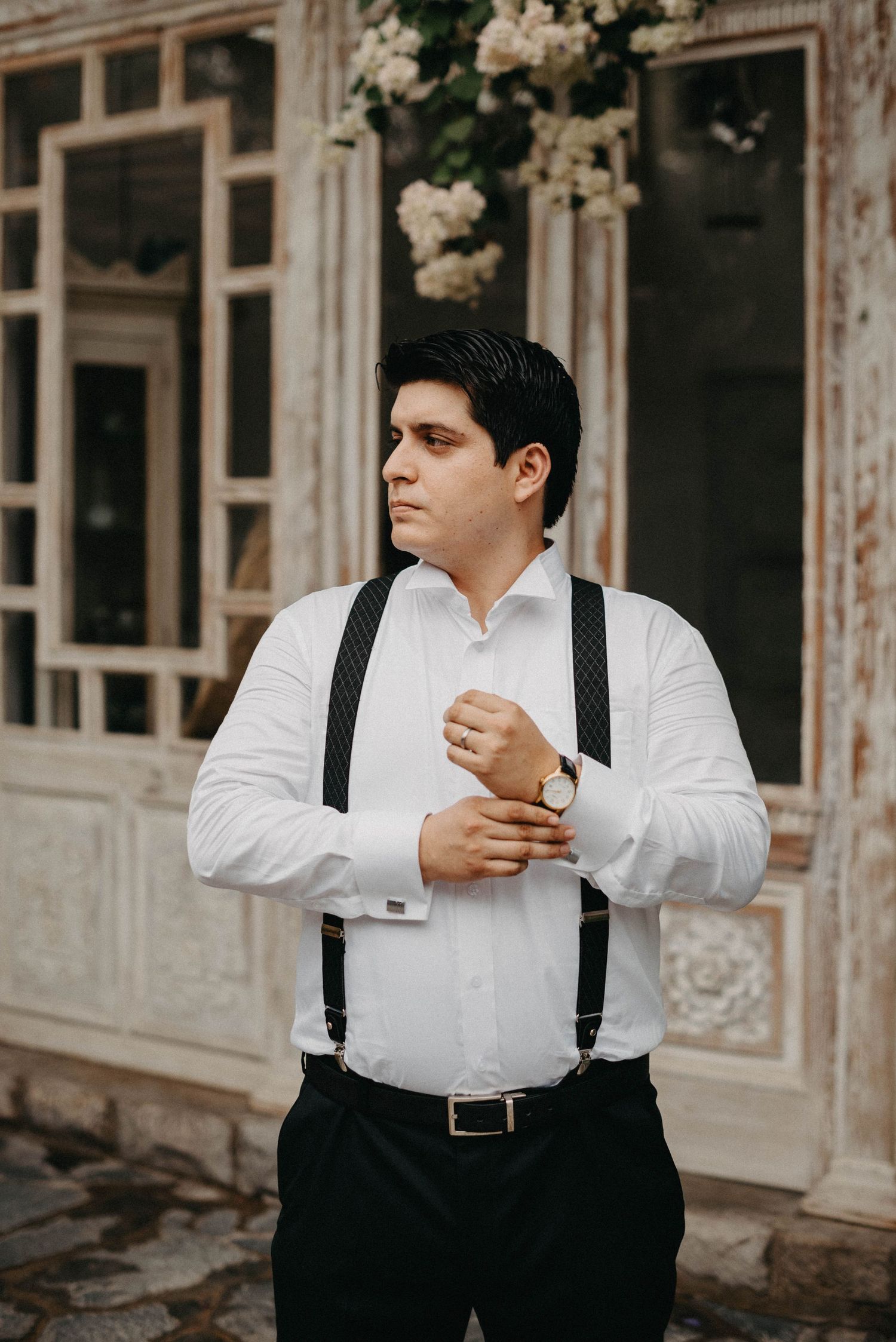 A groom in white shirt and suspenders poses elegantly against a vintage building facade with blooming flowers.