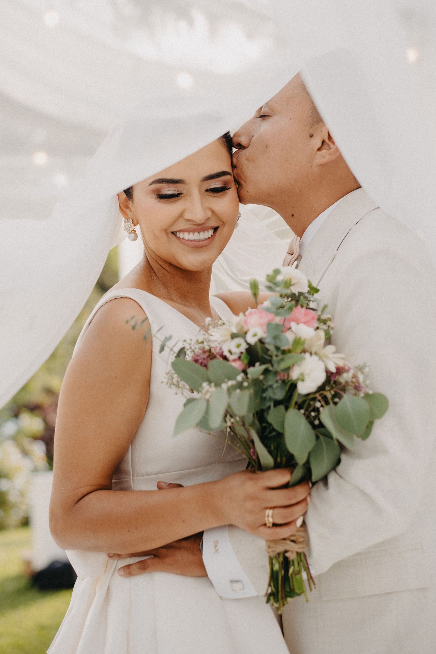 A romantic wedding moment captured outdoors as a couple embraces while holding a soft pink and eucalyptus bouquet.