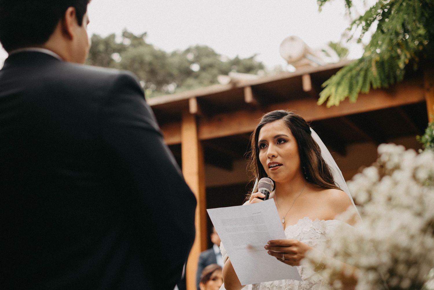 A person in a bridal gown reads from papers during an outdoor wedding ceremony.
