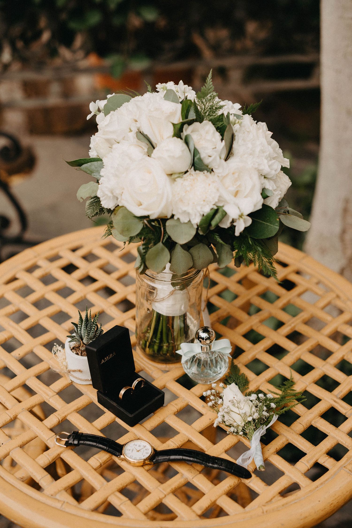 Wedding details including white floral bouquet, ring box, watch and perfume bottle arranged on lattice table.