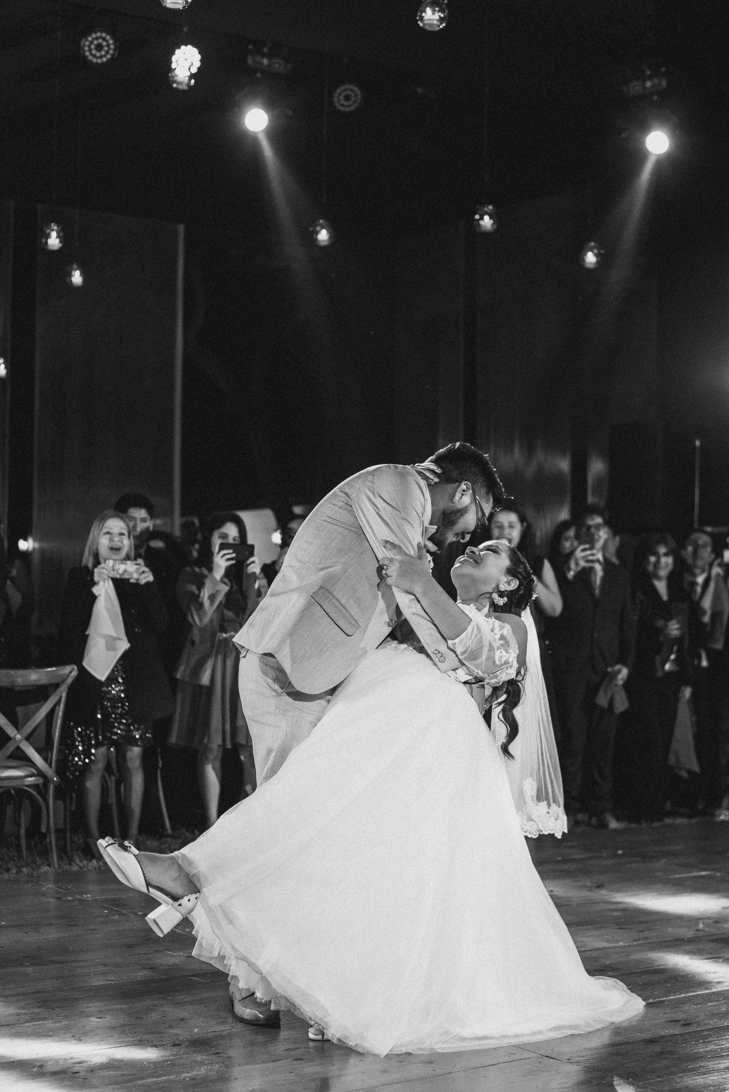 Dramatic black and white photo of couple sharing first dance with groom dipping bride on illuminated dance floor.