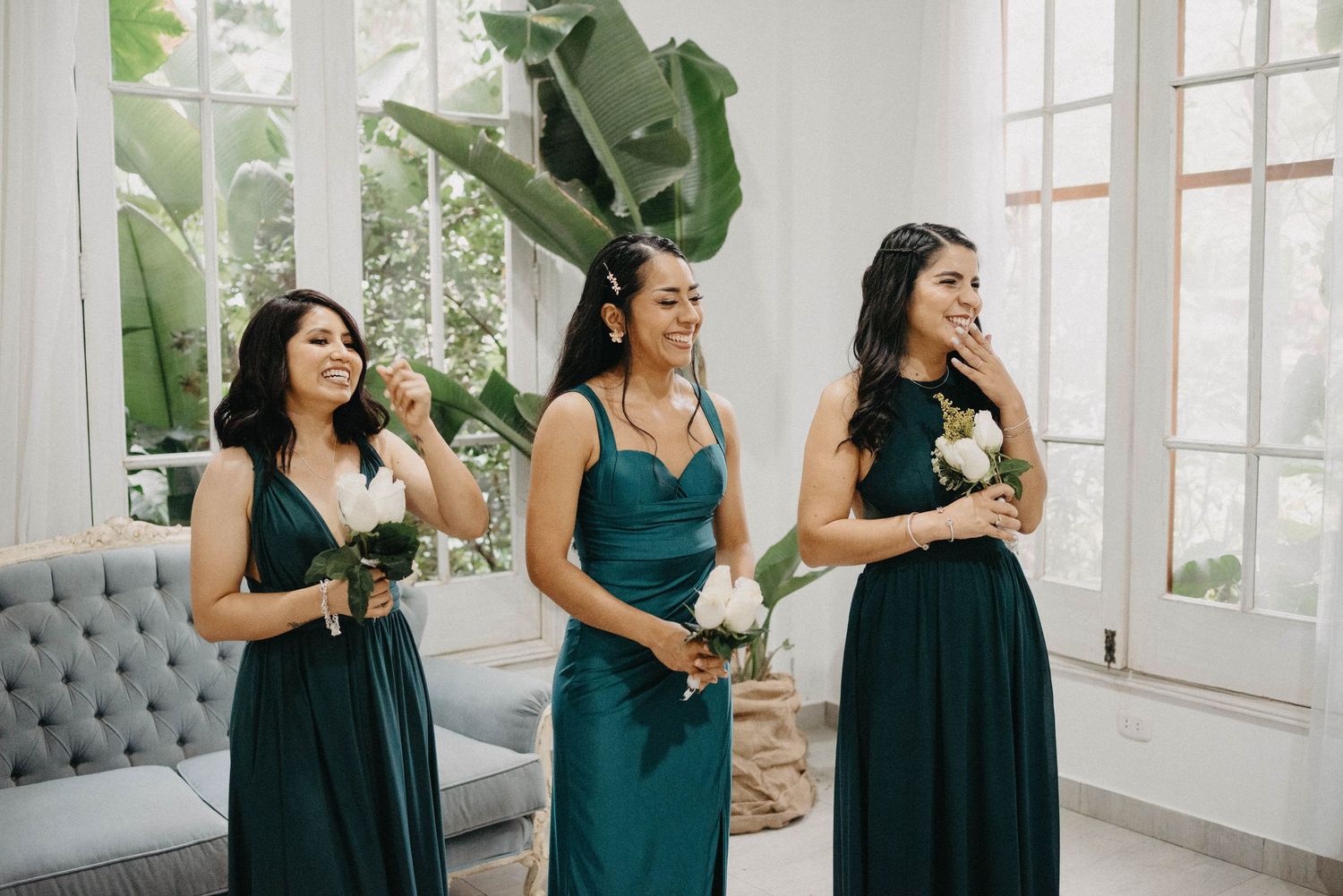 Three bridesmaids in elegant teal gowns laughing together in a bright sunlit room with large plants.