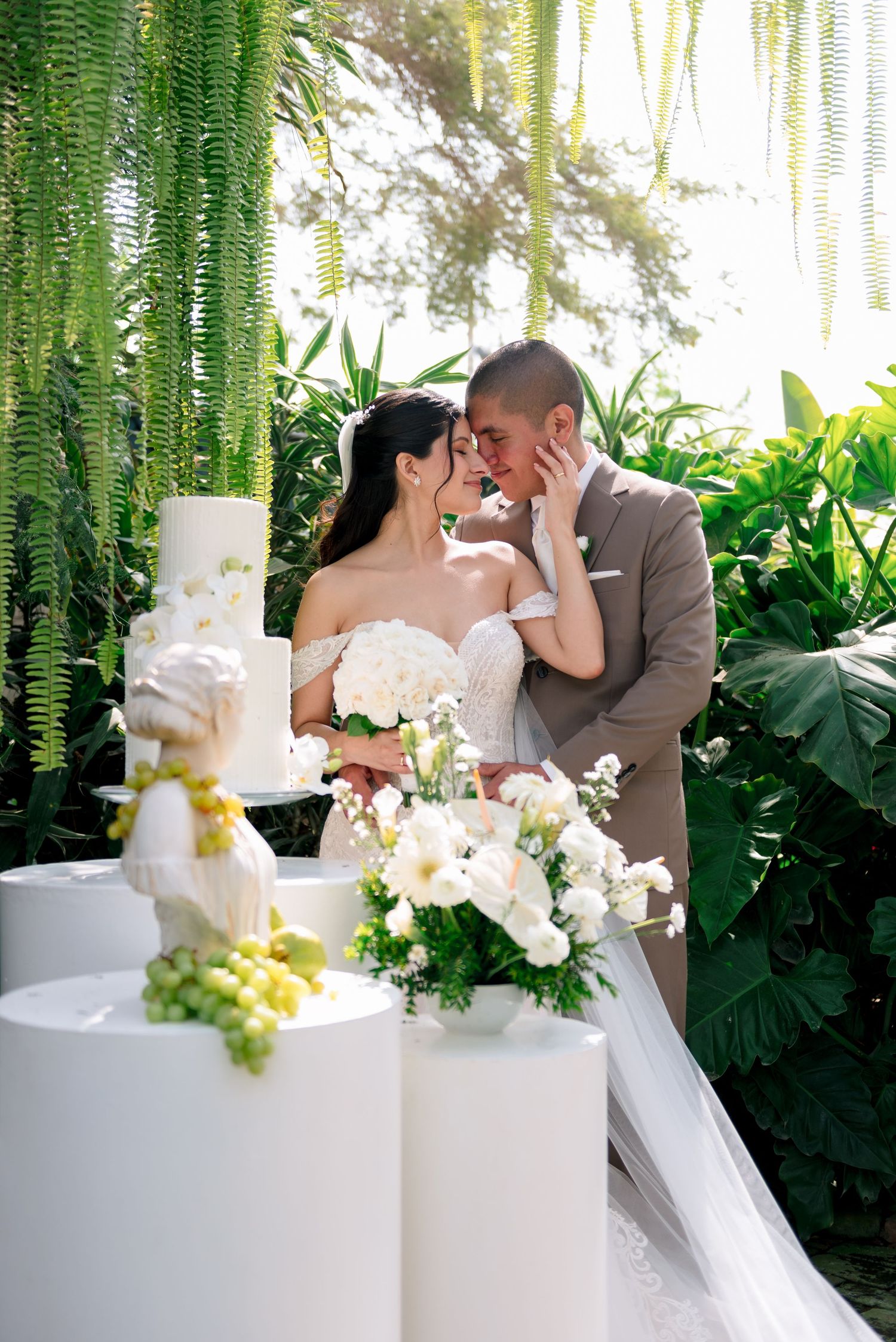 A romantic wedding scene with a white tiered cake adorned with flowers in a lush tropical garden setting.
