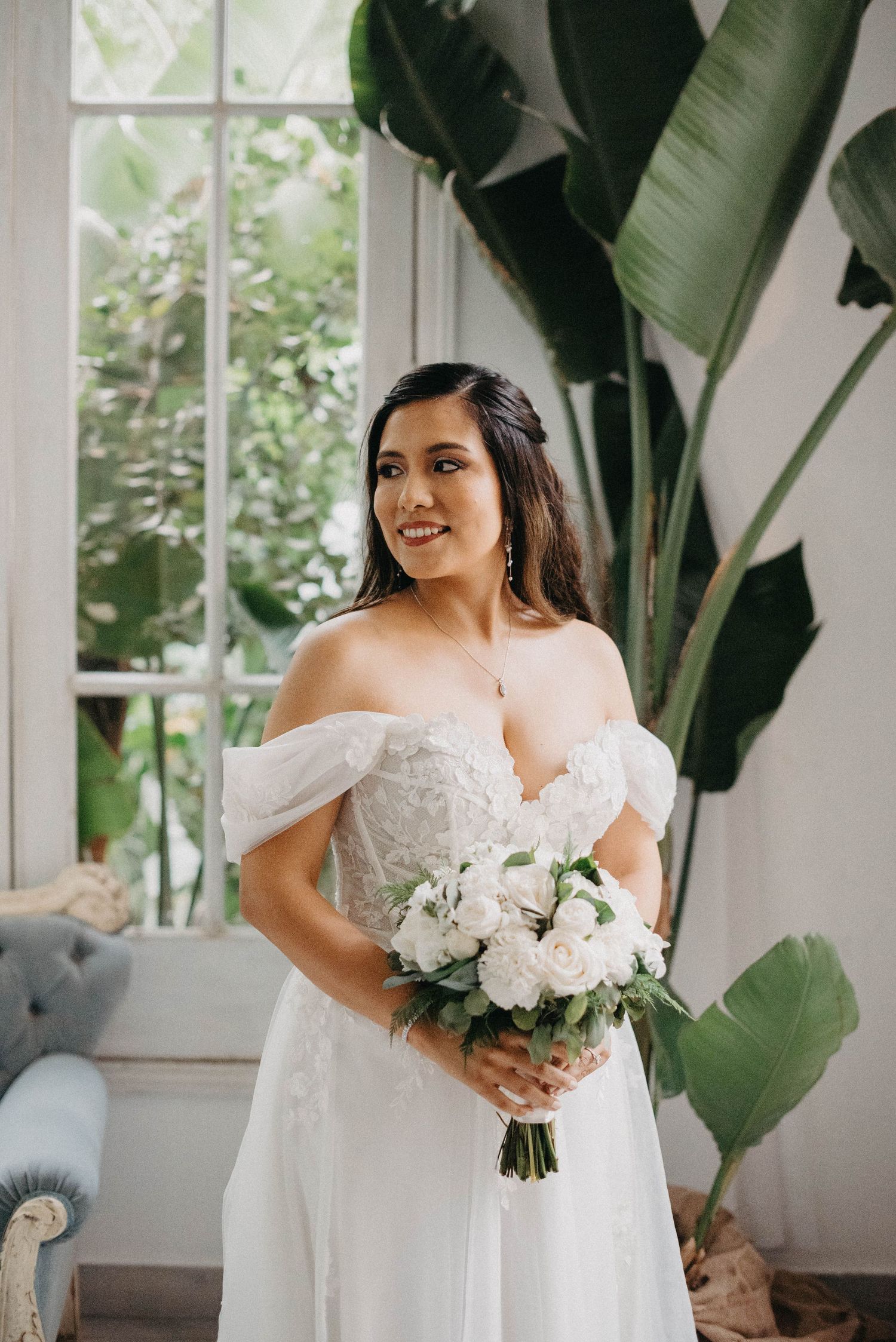 A beautiful bride in an off-shoulder white gown holds a bouquet of white flowers against a backdrop of tropical plants.