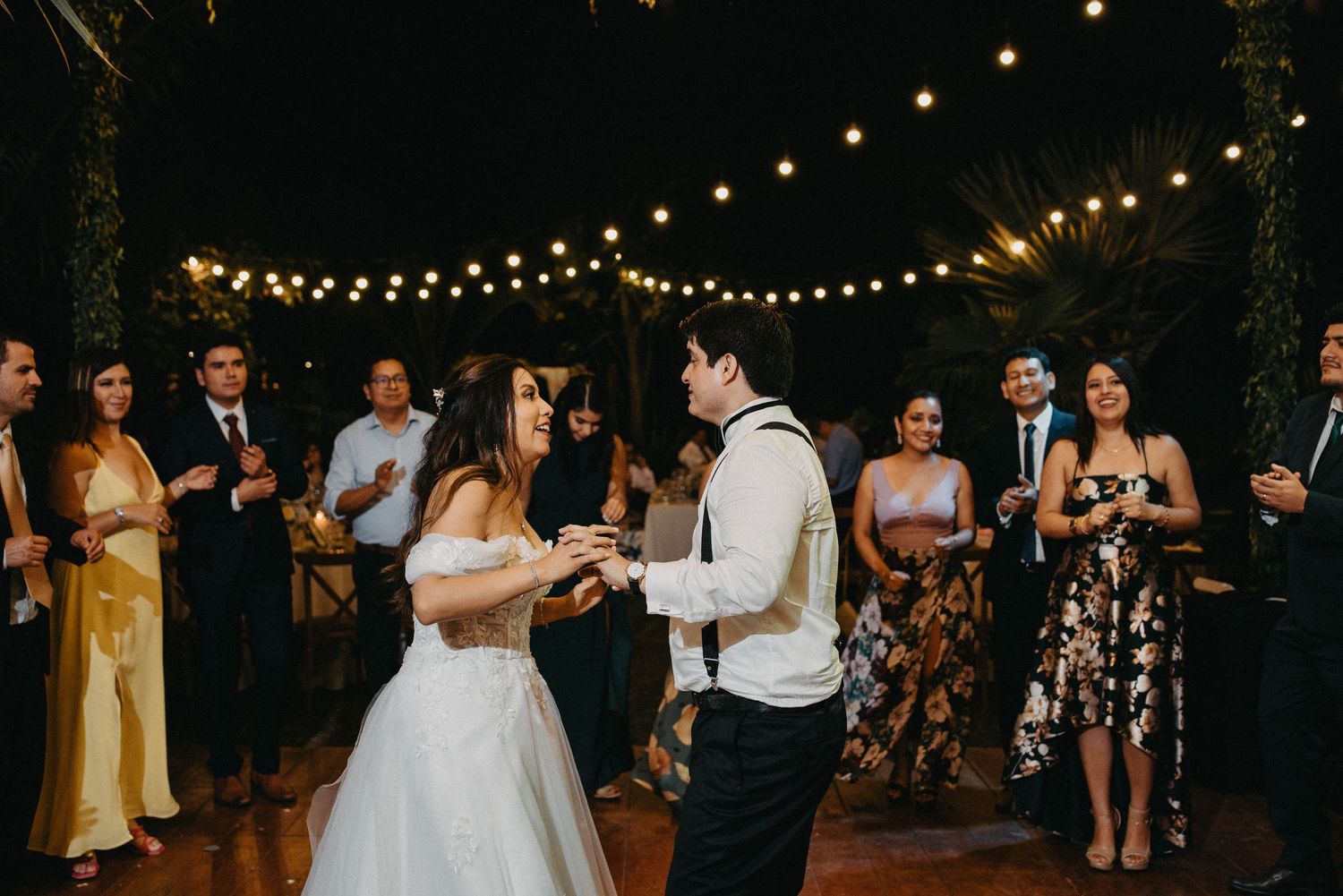 A romantic evening wedding reception under string lights with guests dancing on an outdoor patio.