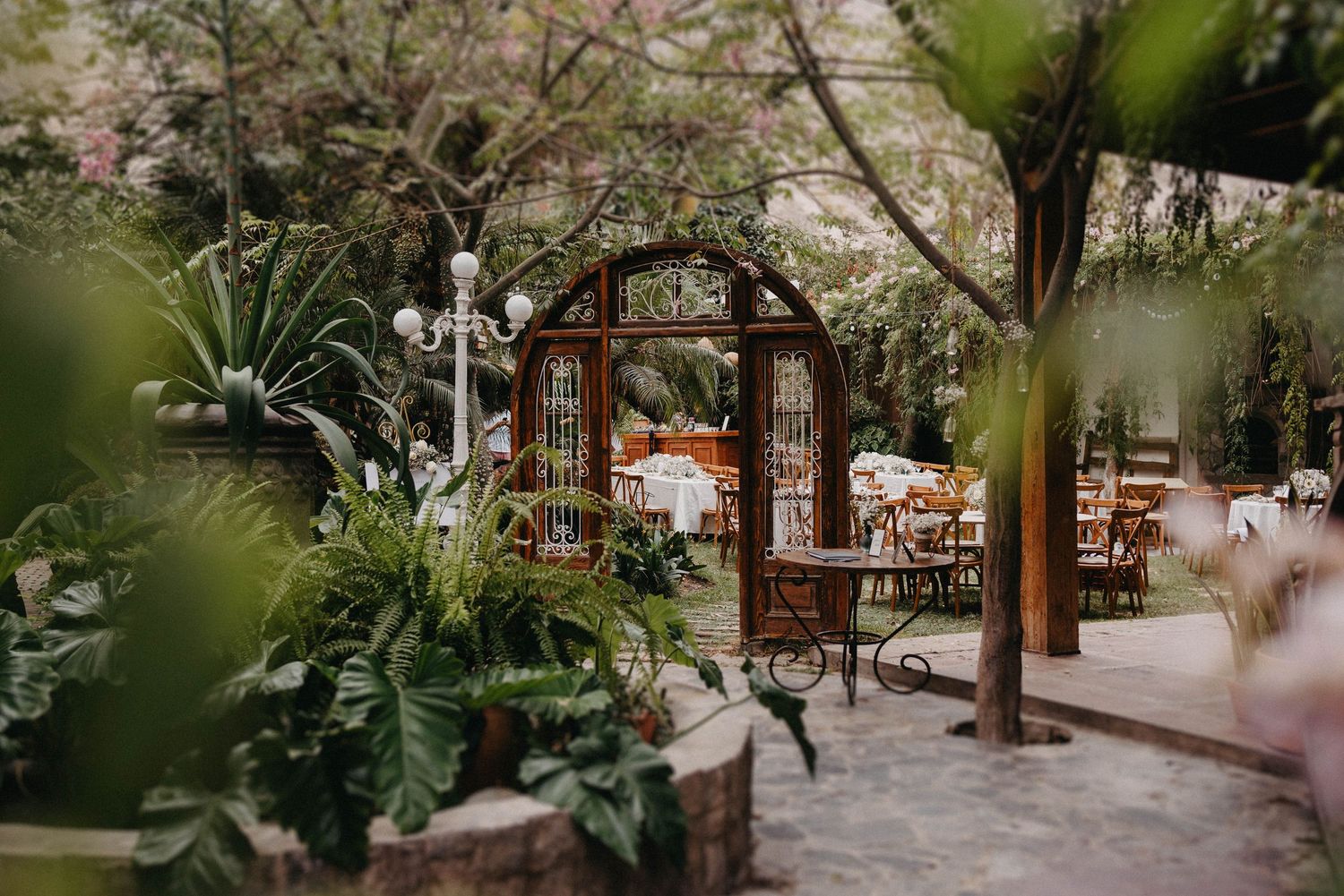 A rustic wooden garden arch leads to an outdoor dining area surrounded by lush tropical plants and greenery.