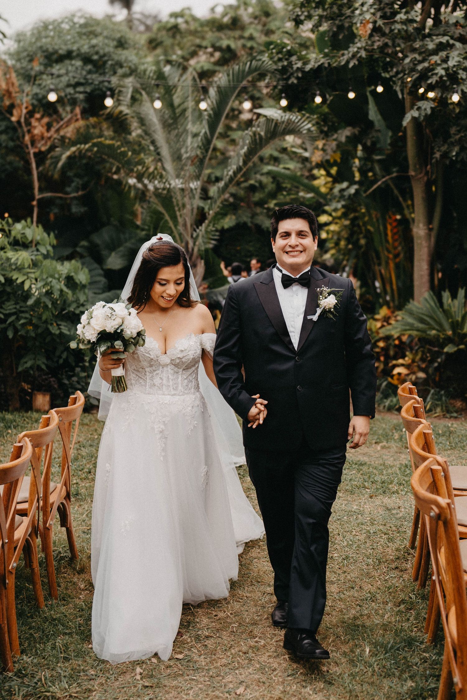 Newlywed couple walking down outdoor garden aisle with string lights and wooden chairs at tropical wedding ceremony.