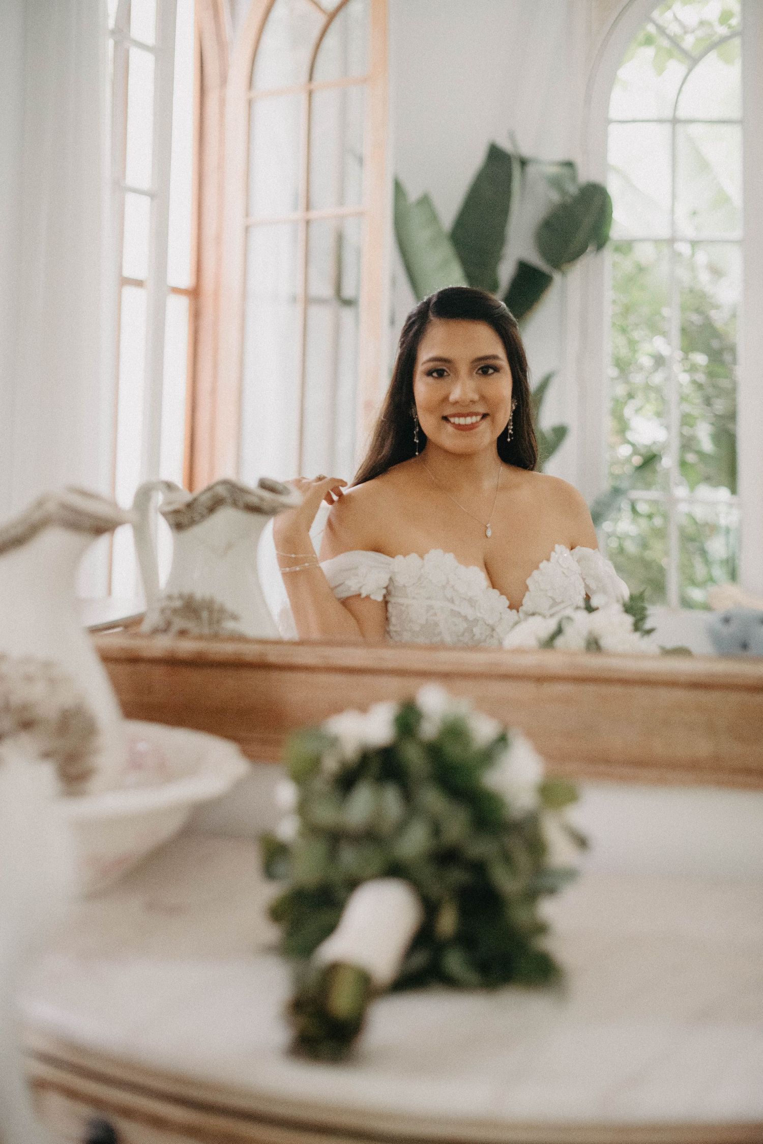 A bride in an off-shoulder wedding gown smiles while sitting at a vanity table with elegant white window treatments.