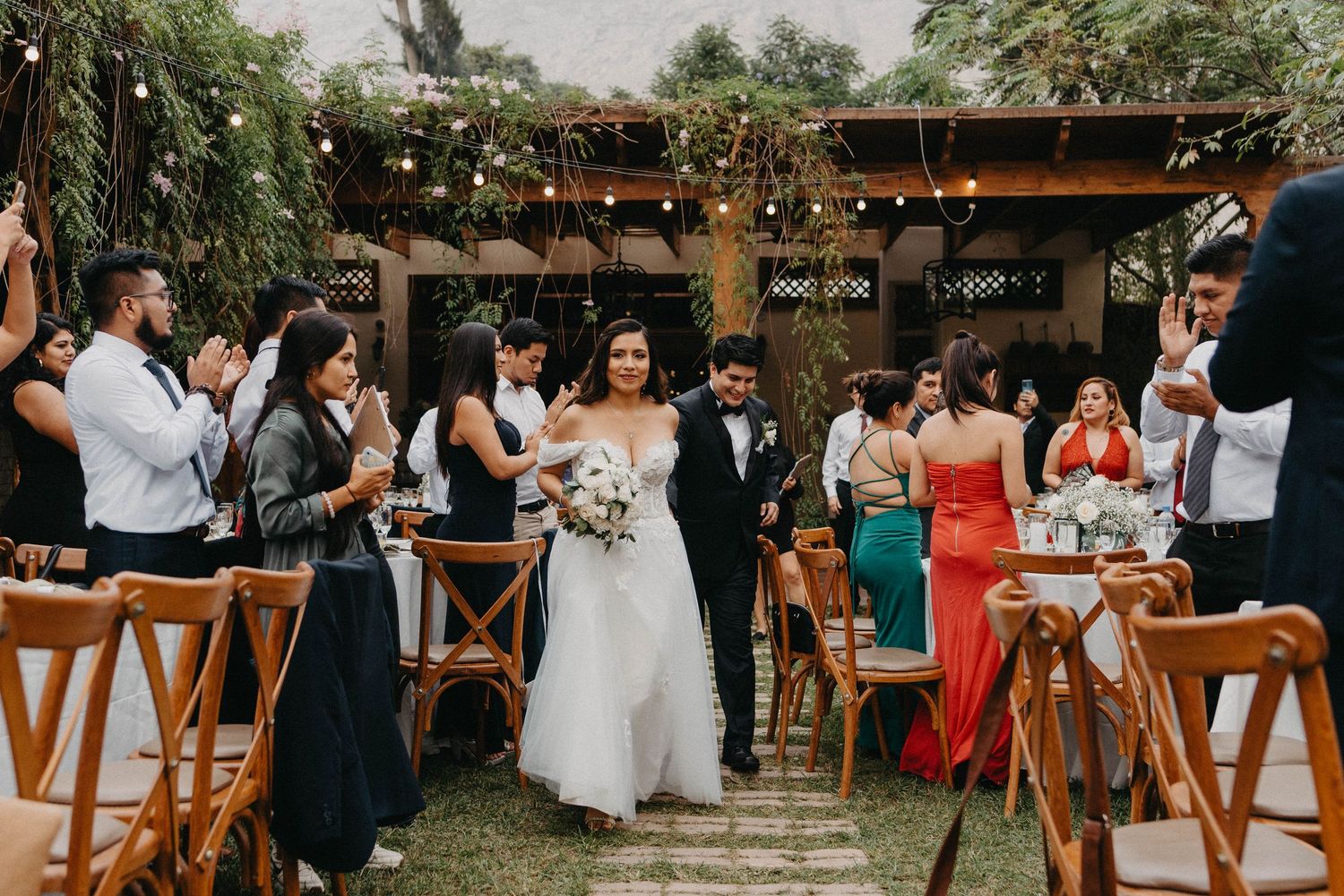 Wedding reception guests seated at long tables under string lights in tropical garden setting.