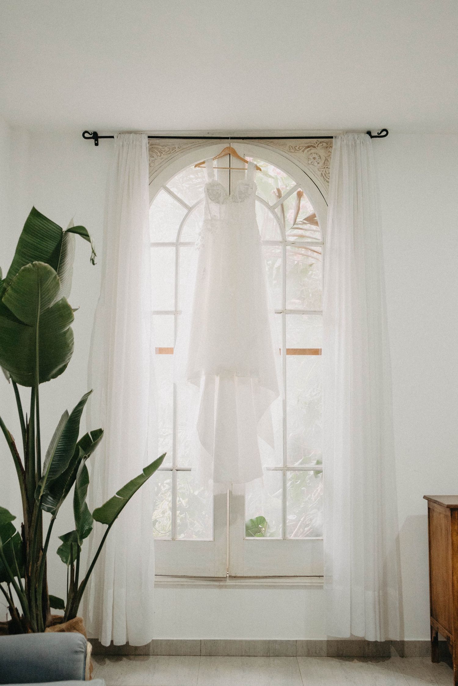 White wedding dress hanging in front of an arched window with flowing sheer curtains and potted plant.
