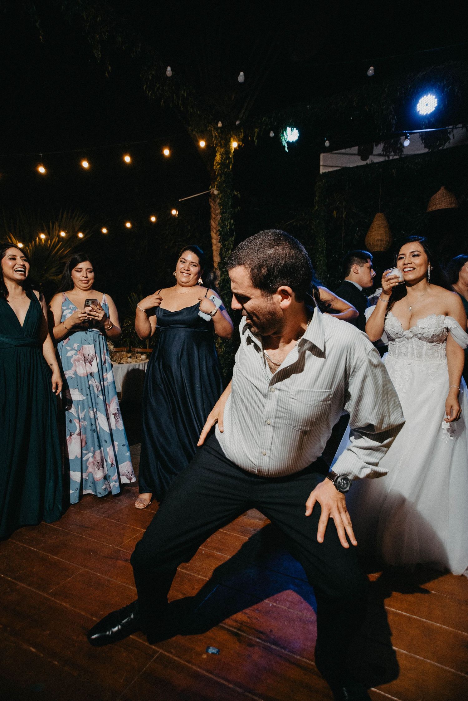 Wedding reception guests perform line dance moves on a dark wooden dance floor at night.