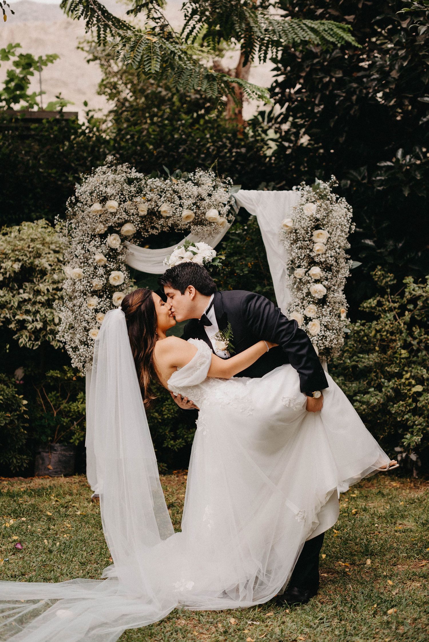 Romantic wedding couple sharing a dip kiss pose against a garden backdrop with white floral arrangements.