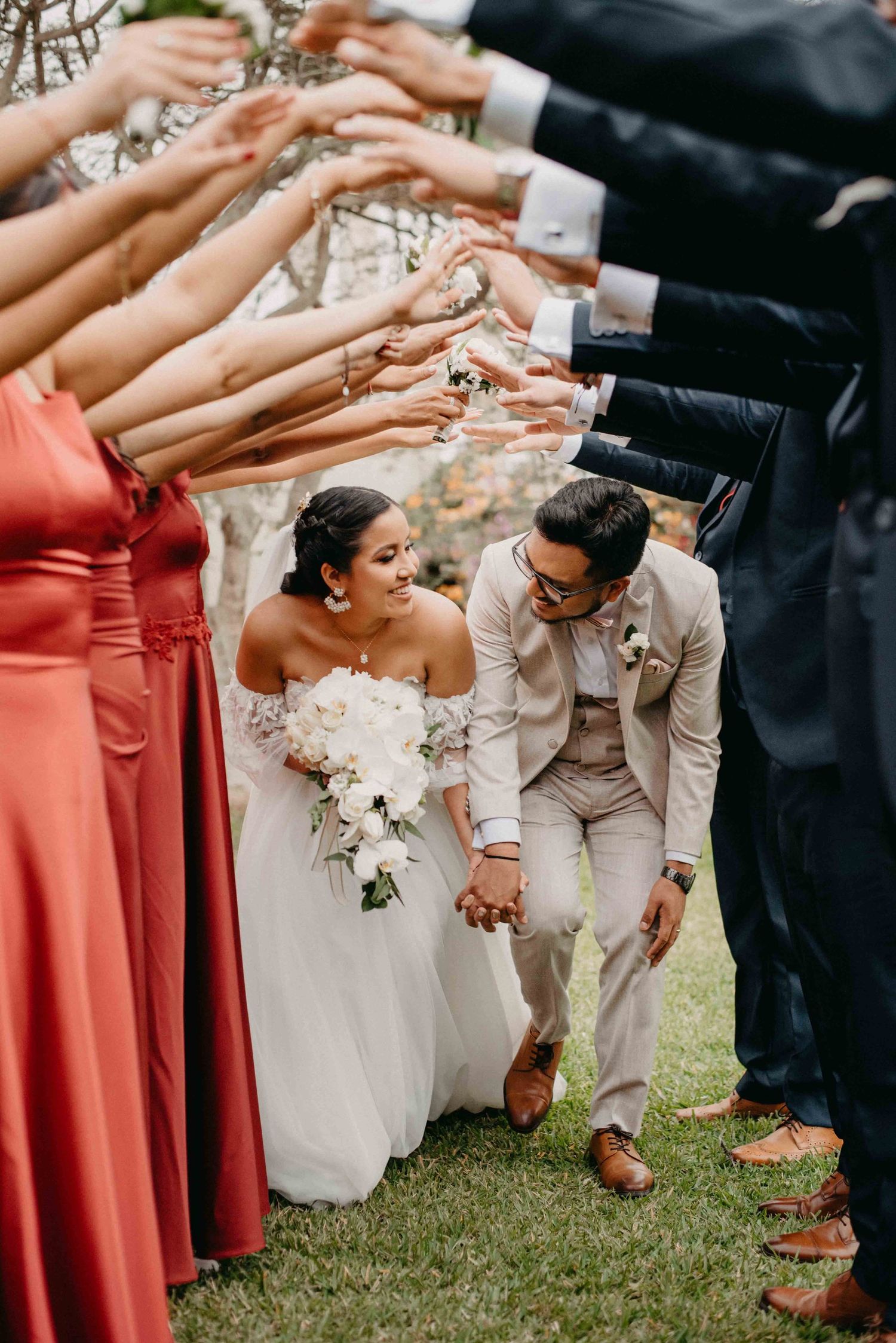 Wedding party in coral dresses and light suits poses together on grass.