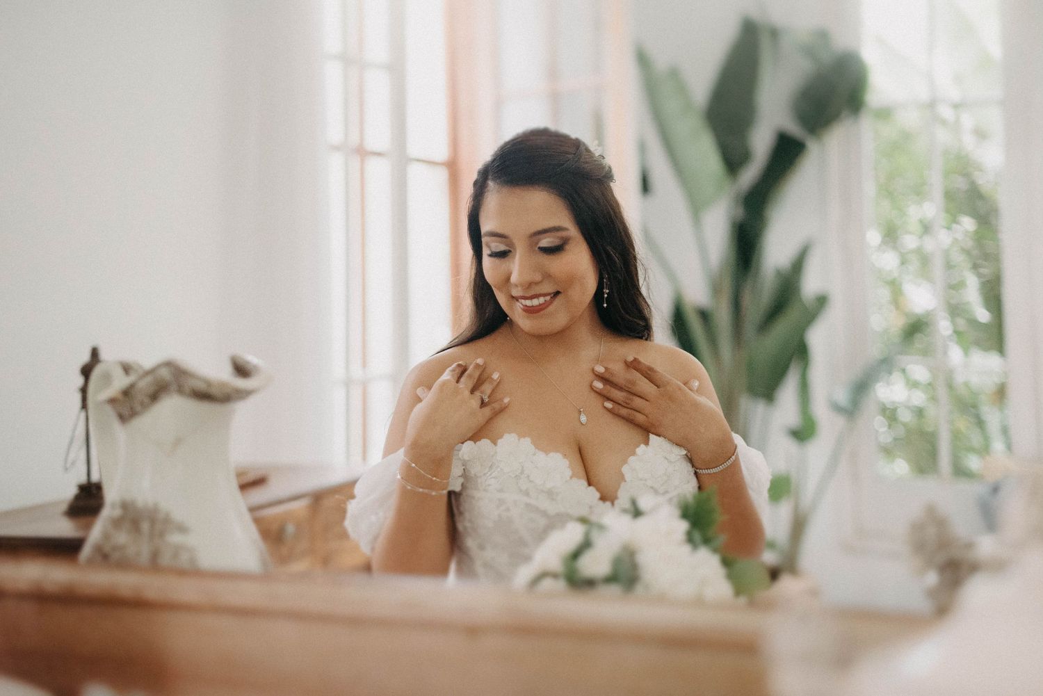 A radiant bride in a strapless dress smiles while touching her necklace near a bright window.