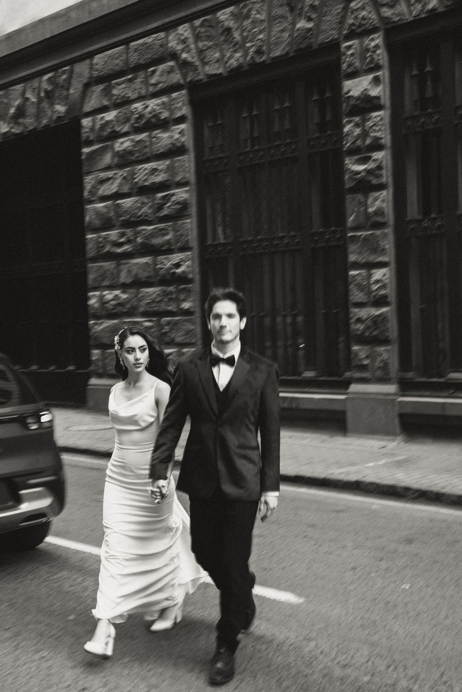A black and white photo of a couple in formal attire walking down a city street beside a stone building.