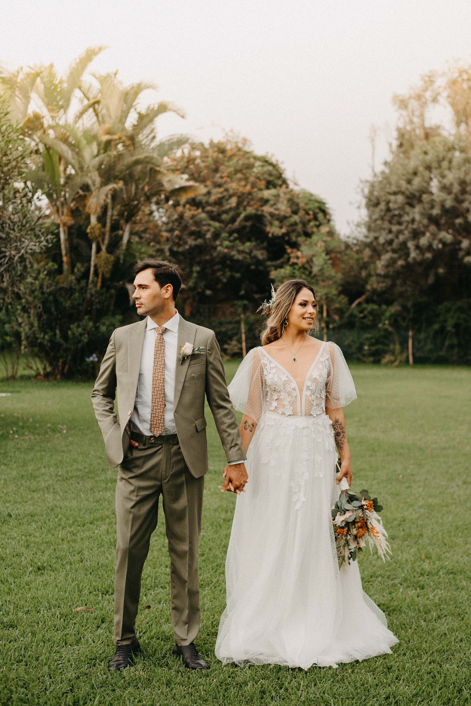 Couple in wedding attire walking through a lush green garden during golden hour photography session.