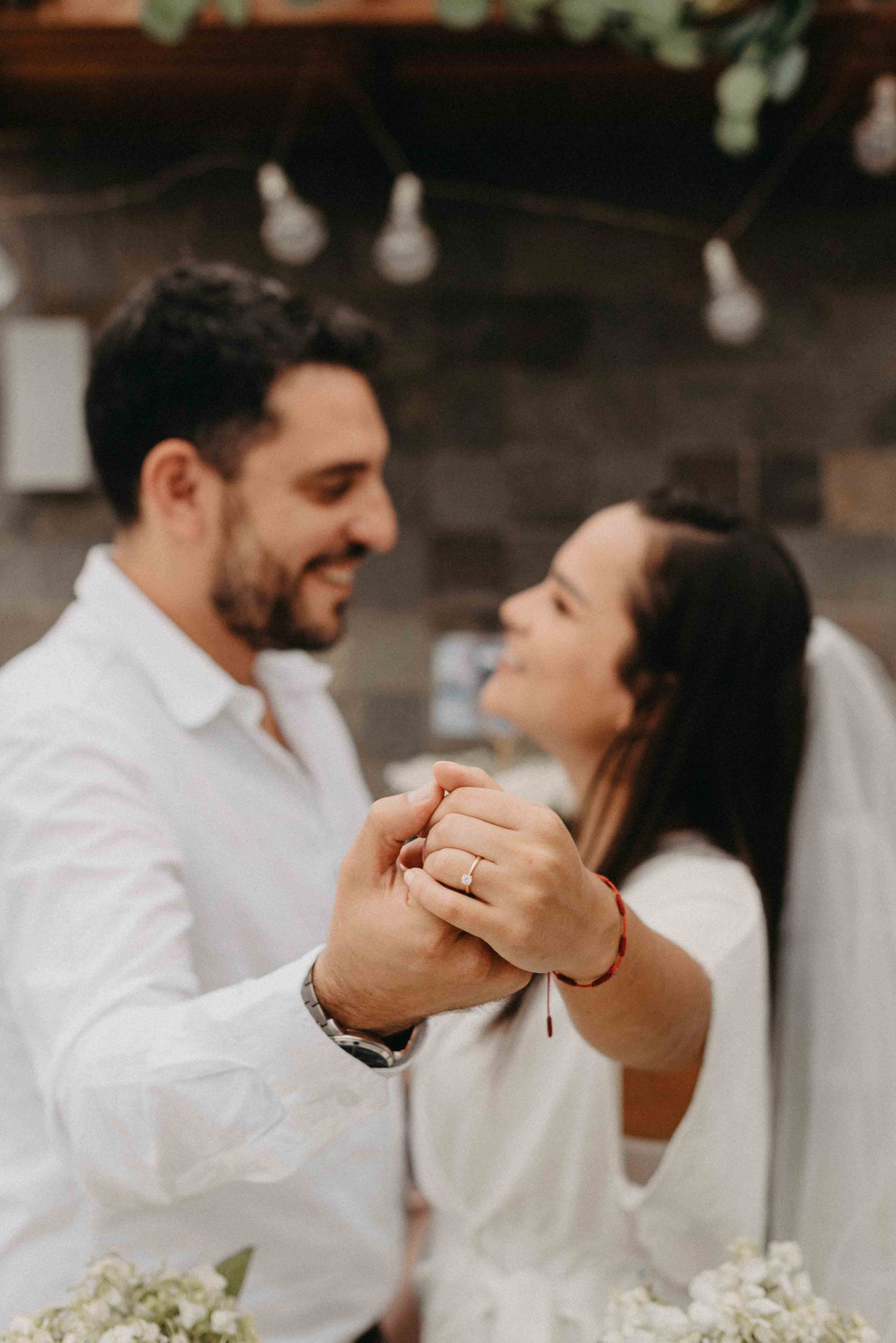 A close-up of two hands clasped together showing an engagement ring with string lights in background.