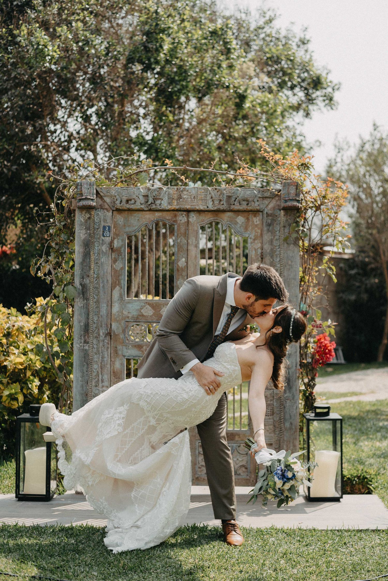 Romantic wedding couple shares passionate dip kiss in garden archway setting.