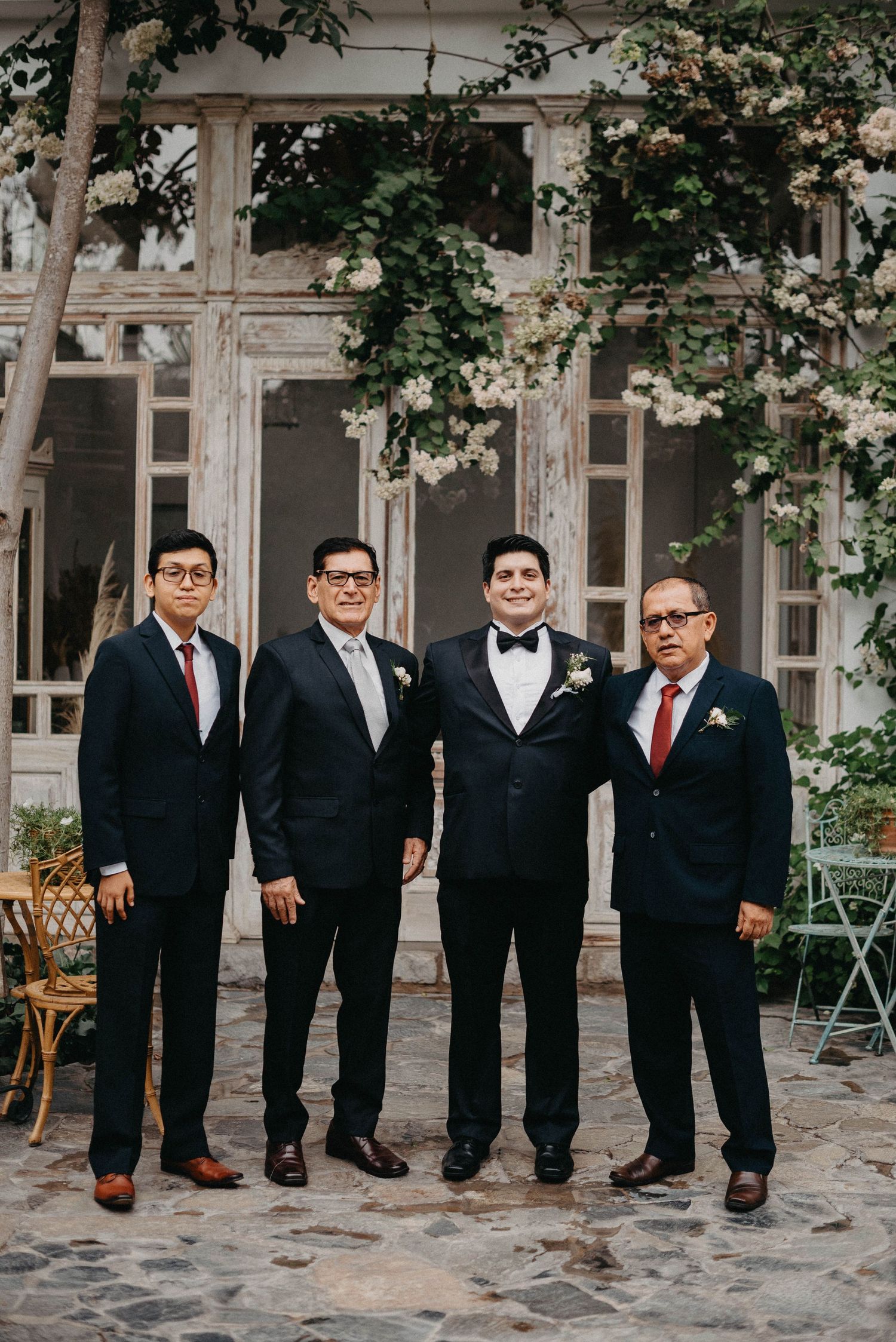 Groomsmen in black suits and bowties pose together at an elegant outdoor wedding venue.