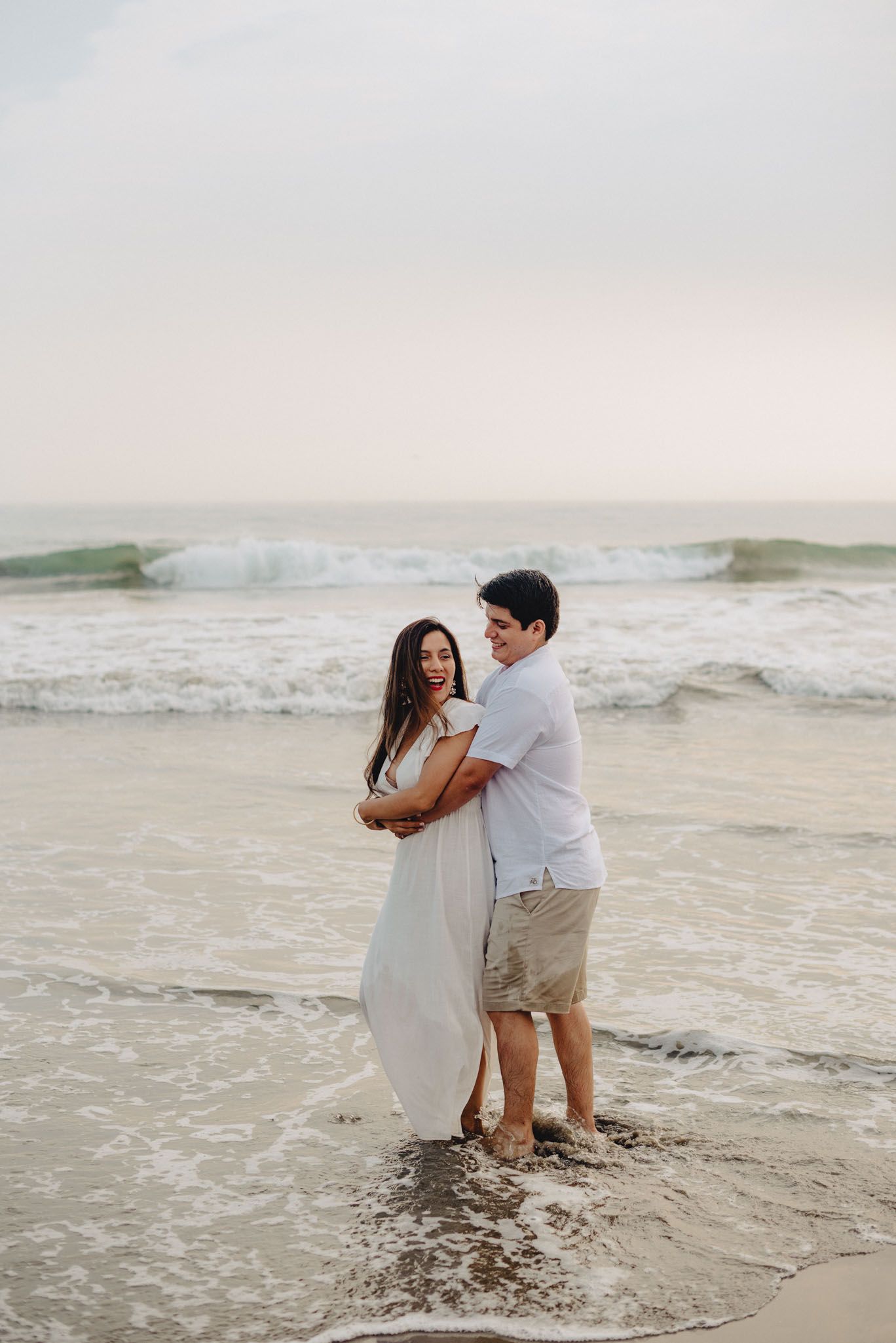 A couple embracing on a sandy beach at sunset with gentle waves rolling in behind them.