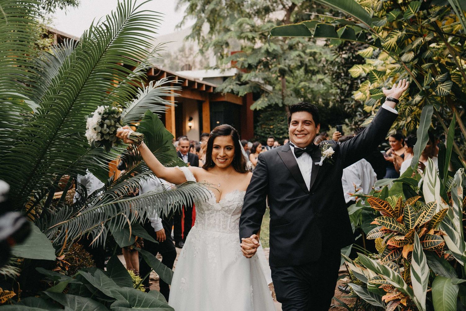 Newlyweds celebrate with raised arms walking through a lush tropical garden setting.