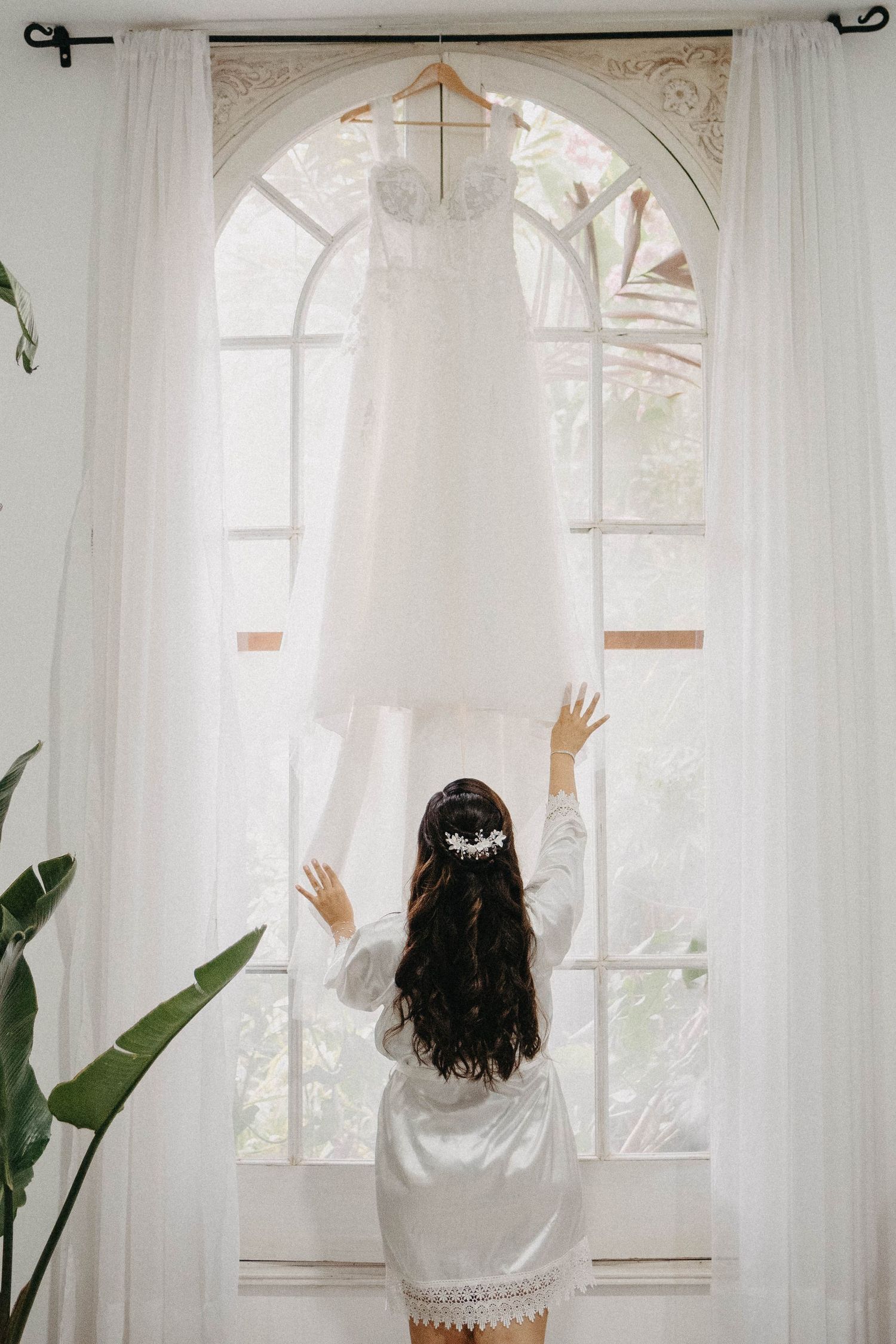 A person in a silk robe reaches up towards a hanging wedding dress by an arched window.