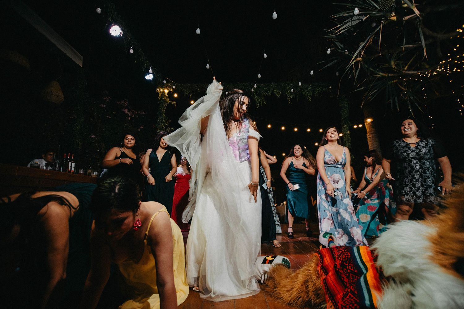 A person in a flowing white wedding dress dances surrounded by guests at a dimly lit reception venue.