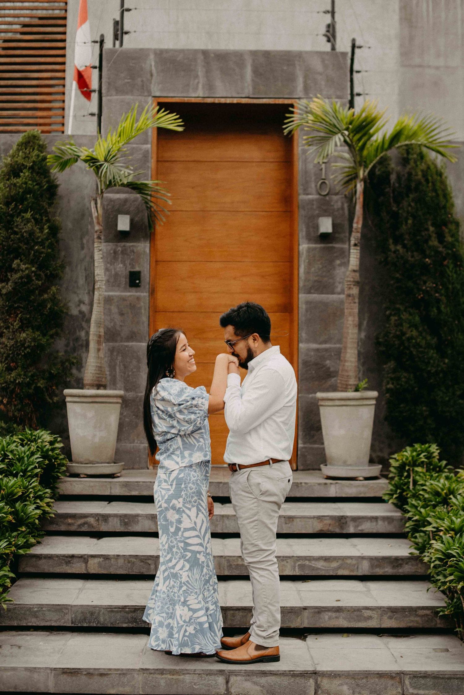 A couple shares a romantic moment on stone steps framed by palm plants and an orange door.