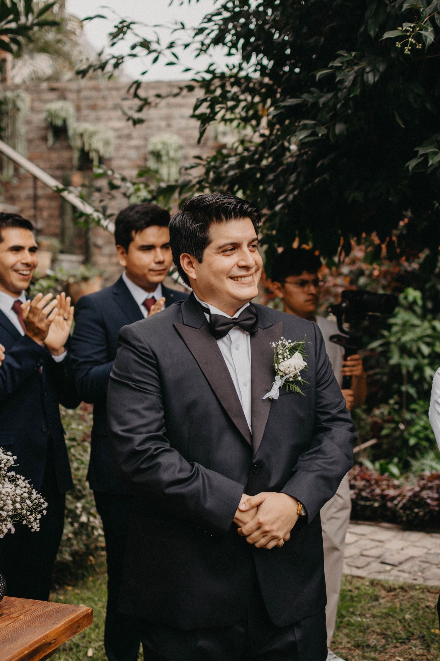 Formal wedding party member in black tuxedo with boutonniere during outdoor garden ceremony.