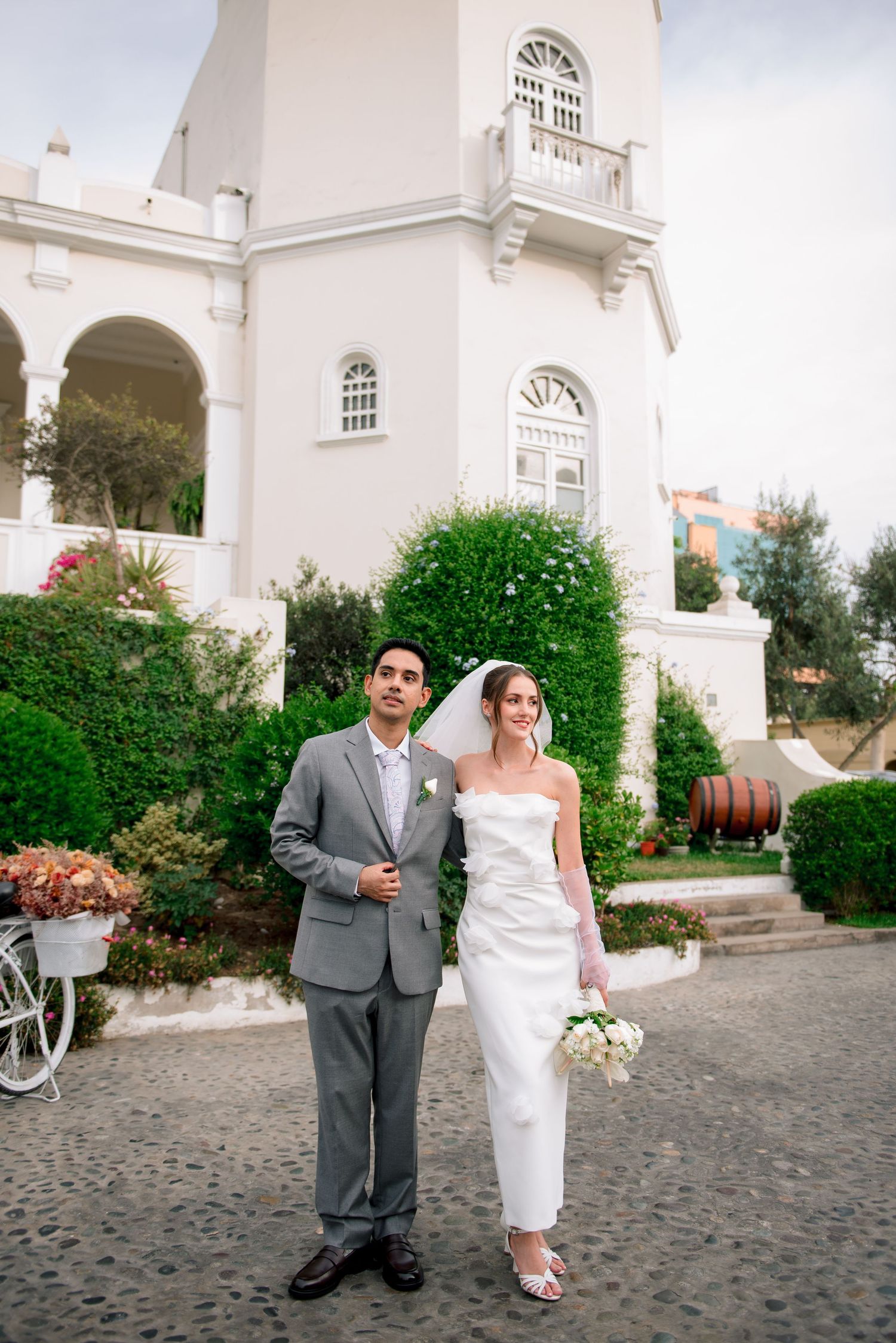 A wedding couple stands in front of a white church with arched windows and lush greenery on a cobblestone path.