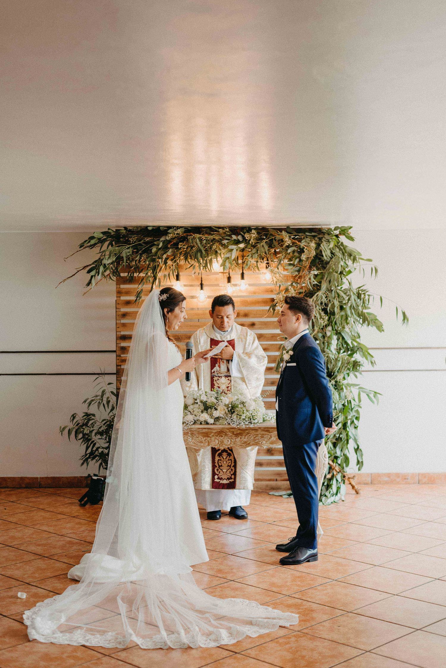 Wedding ceremony under a greenery-adorned arch with officiant and couple exchanging vows.