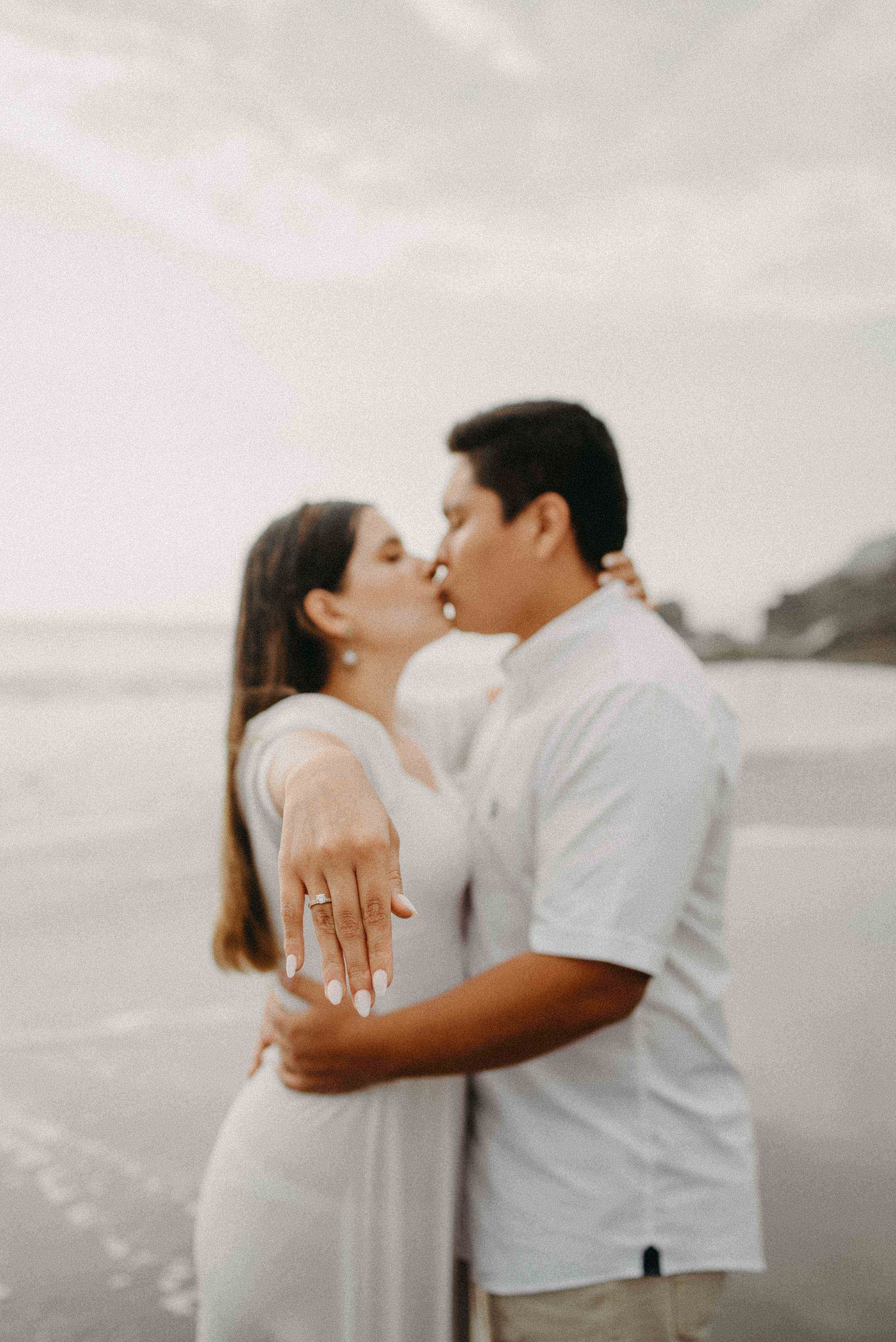 Couple in white embracing on a beach during sunset showing off engagement ring.