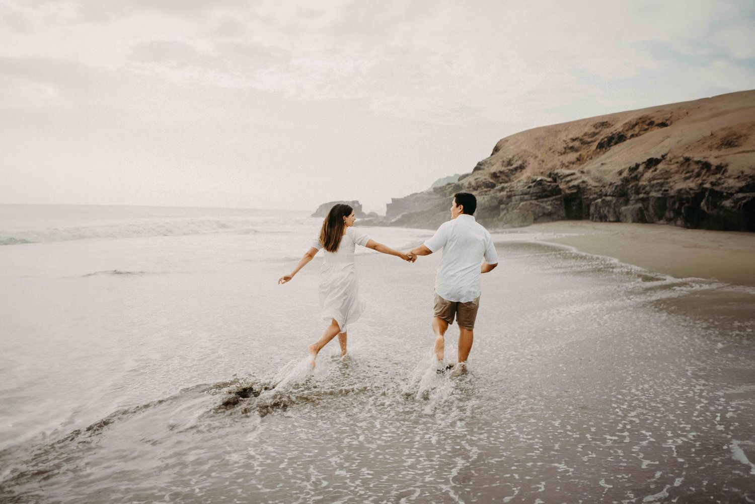 A couple runs playfully along a scenic beach with their dog on their wedding day.