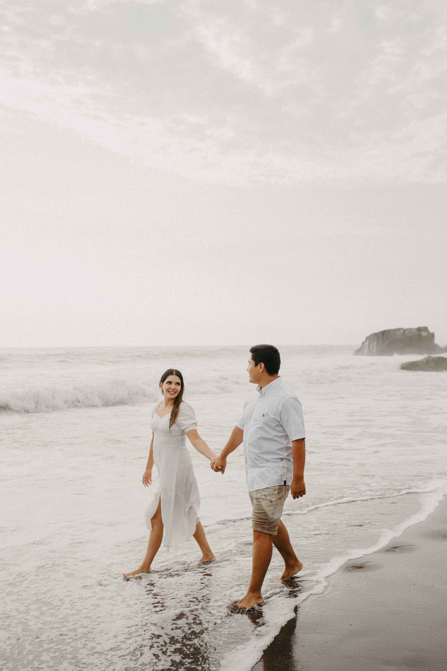 A couple walking hand in hand along a misty beach shoreline with waves crashing in the background.